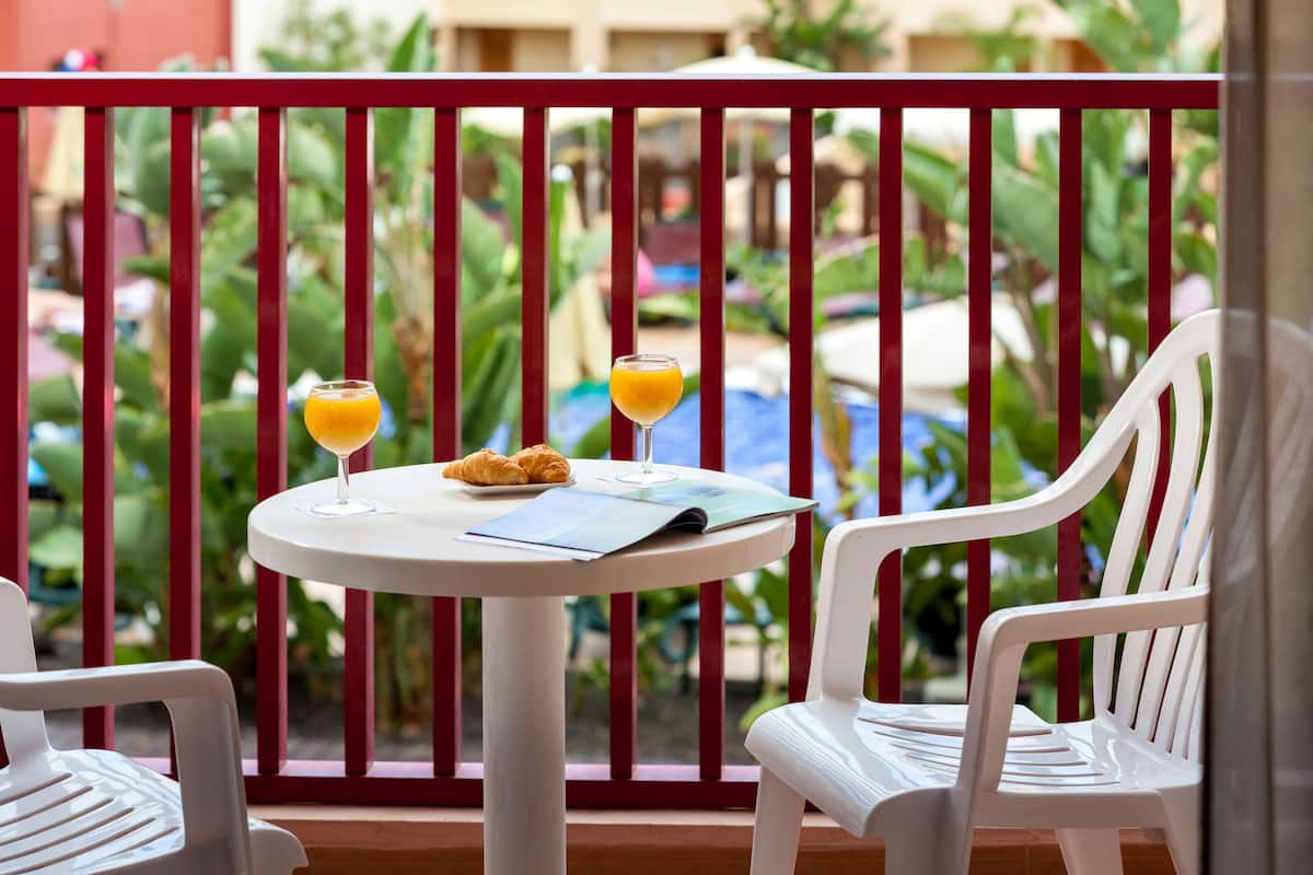 Un balcón con una mesa dispuesta para dos personas, con croissants y vasos de zumo de naranja, con vistas a una piscina y abundante vegetación.
