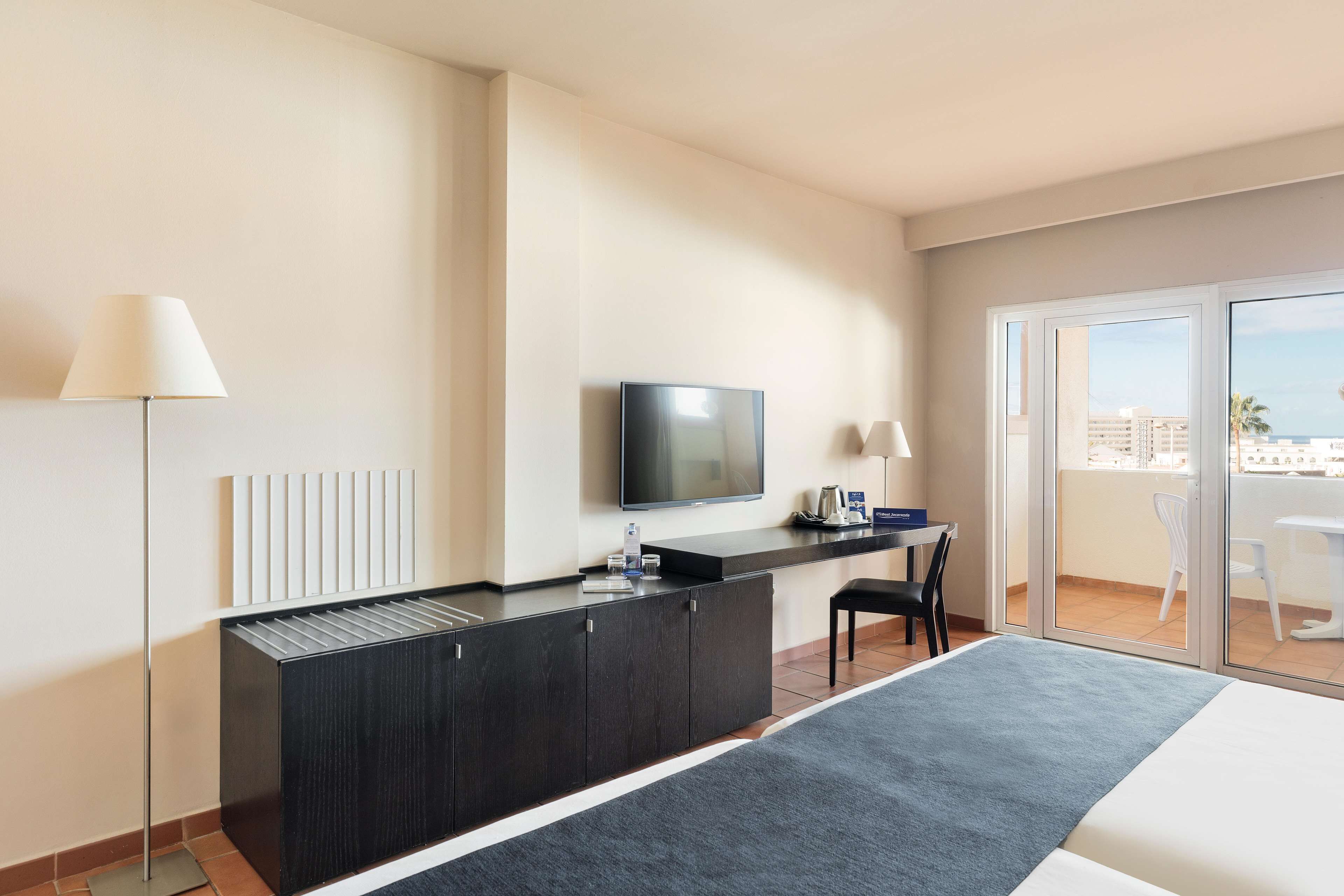Habitación de hotel con cama, escritorio con televisor, silla, lámpara y vista a la ciudad desde la ventana.