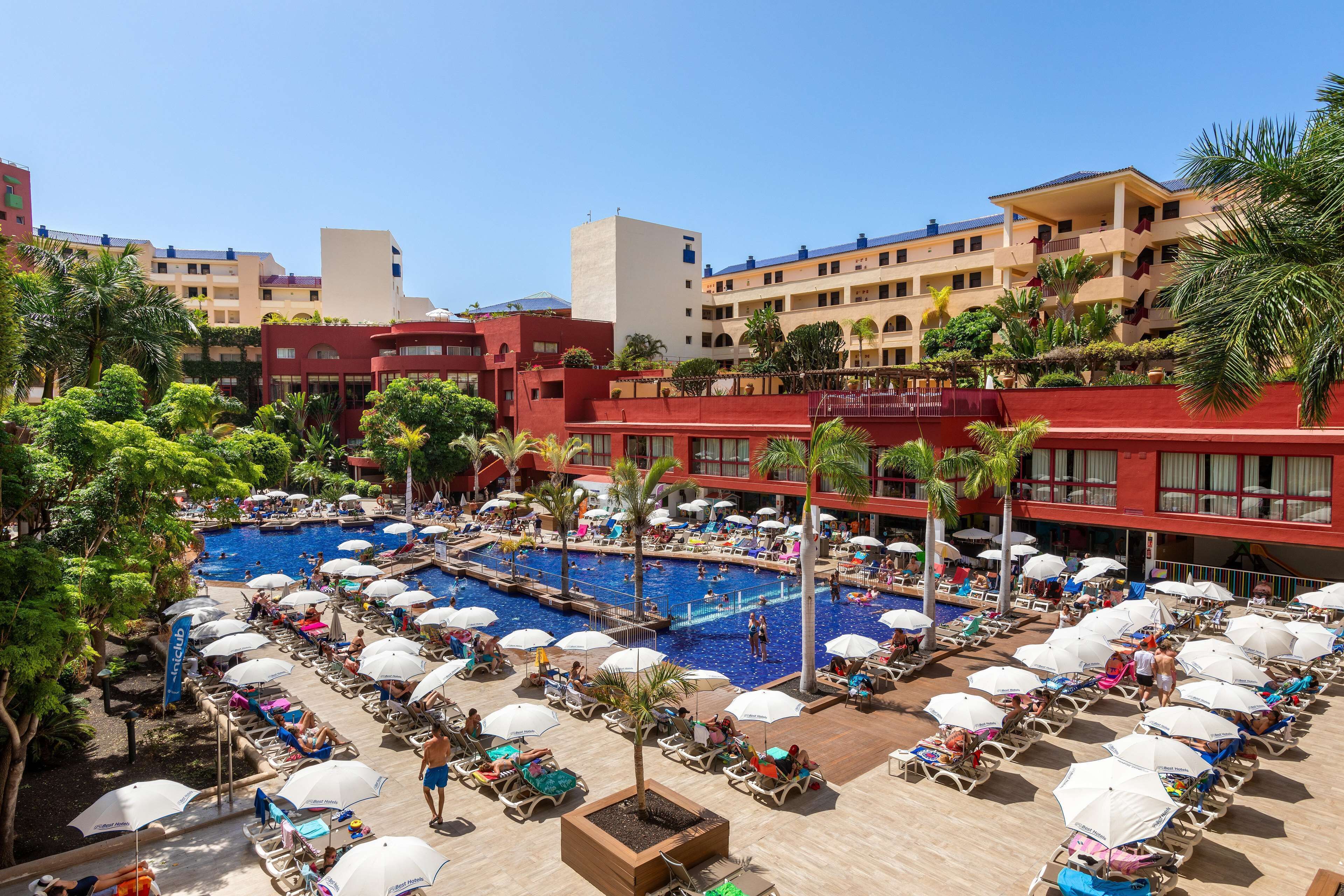 Ein Resort mit einem großen Schwimmbad, umgeben von Liegestühlen und Sonnenschirmen, mehreren Gebäuden mit Balkonen und üppiger Vegetation.