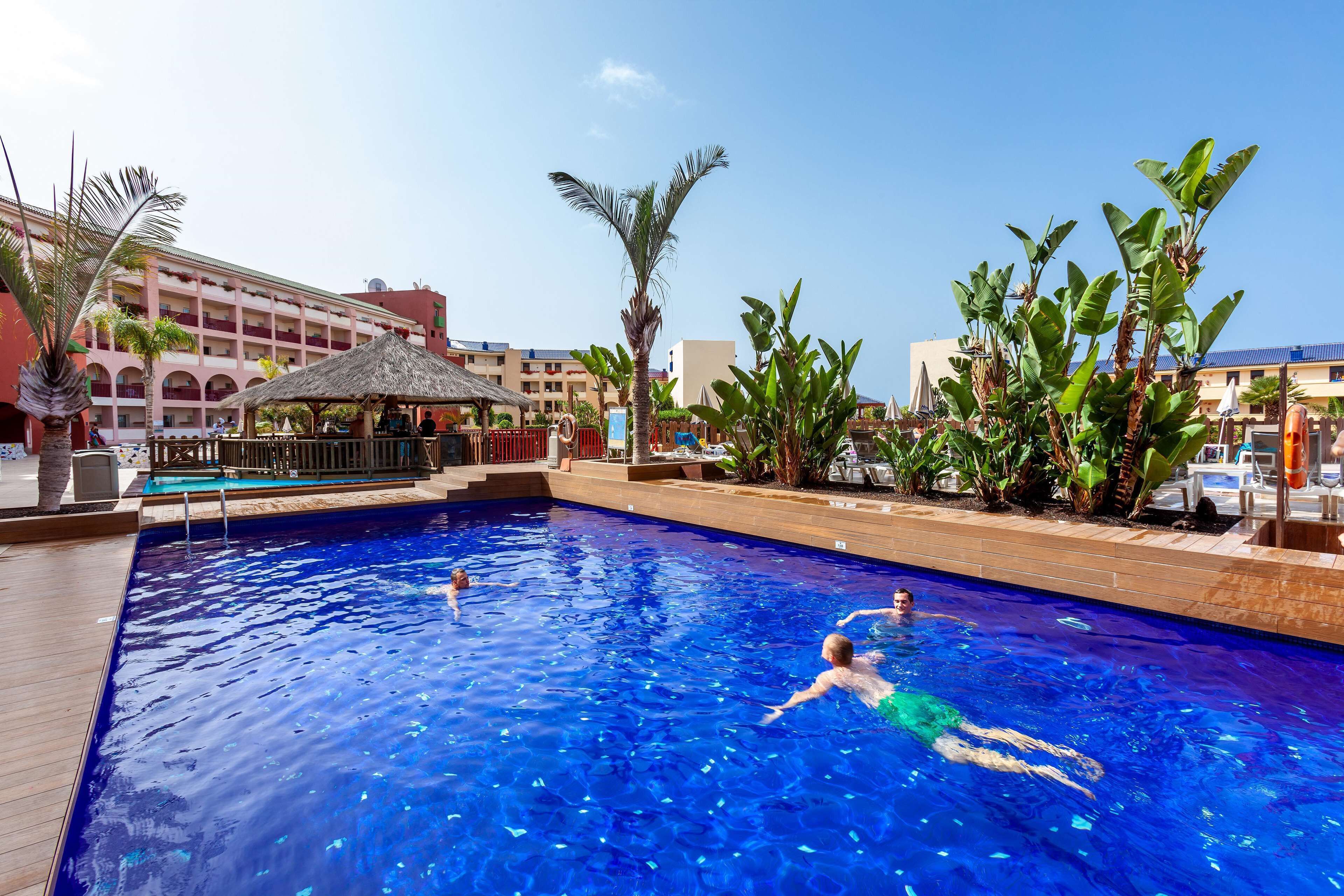 A swimming pool with people swimming, surrounded by palm trees and a building with balconies.