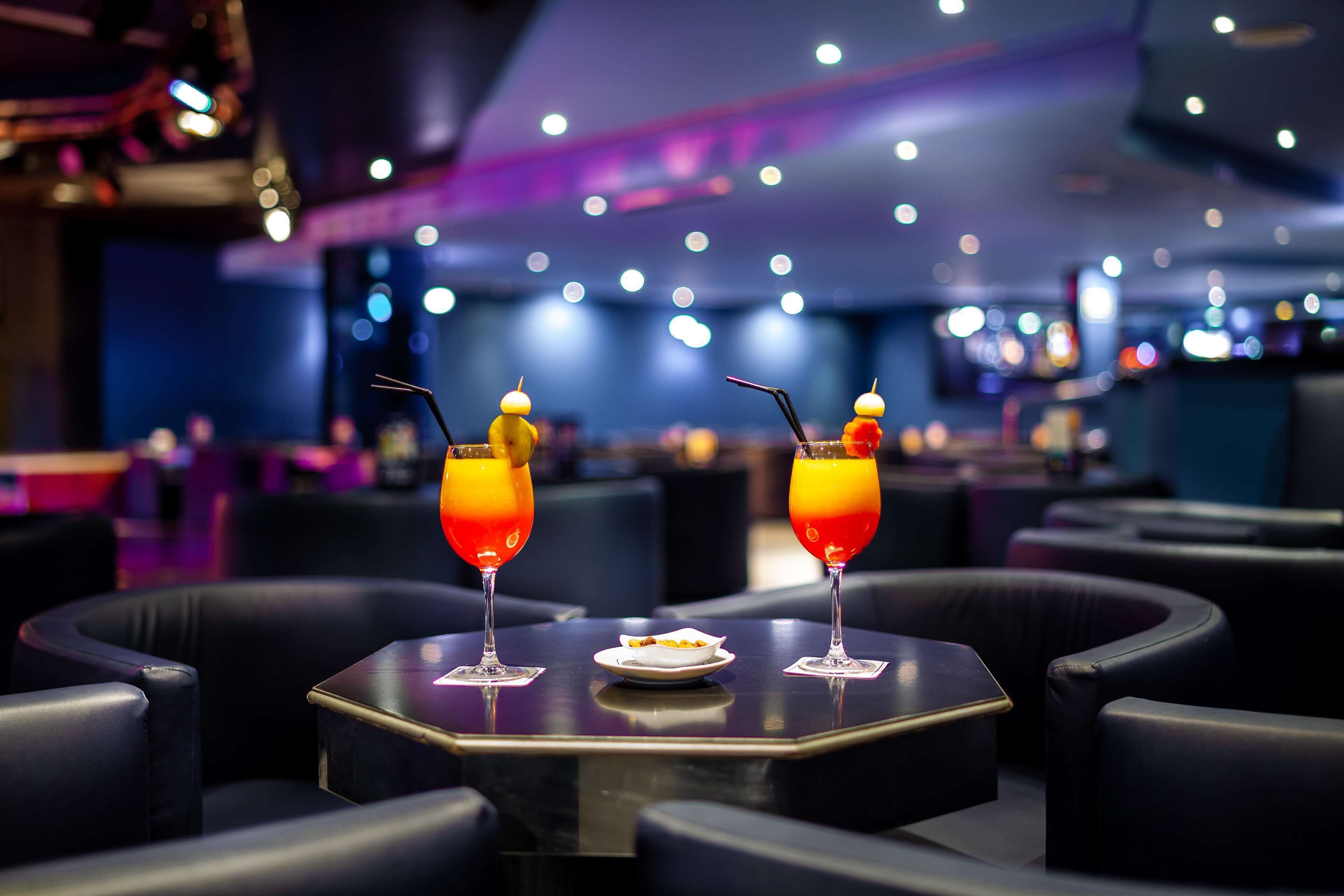 Un coin bar avec deux tables, chacune agrémentée d’un verre de jus d’orange et d’une petite assiette de nourriture. L’ambiance est rehaussée par des fauteuils en cuir foncé et un plafond moderne doté de luminaires.