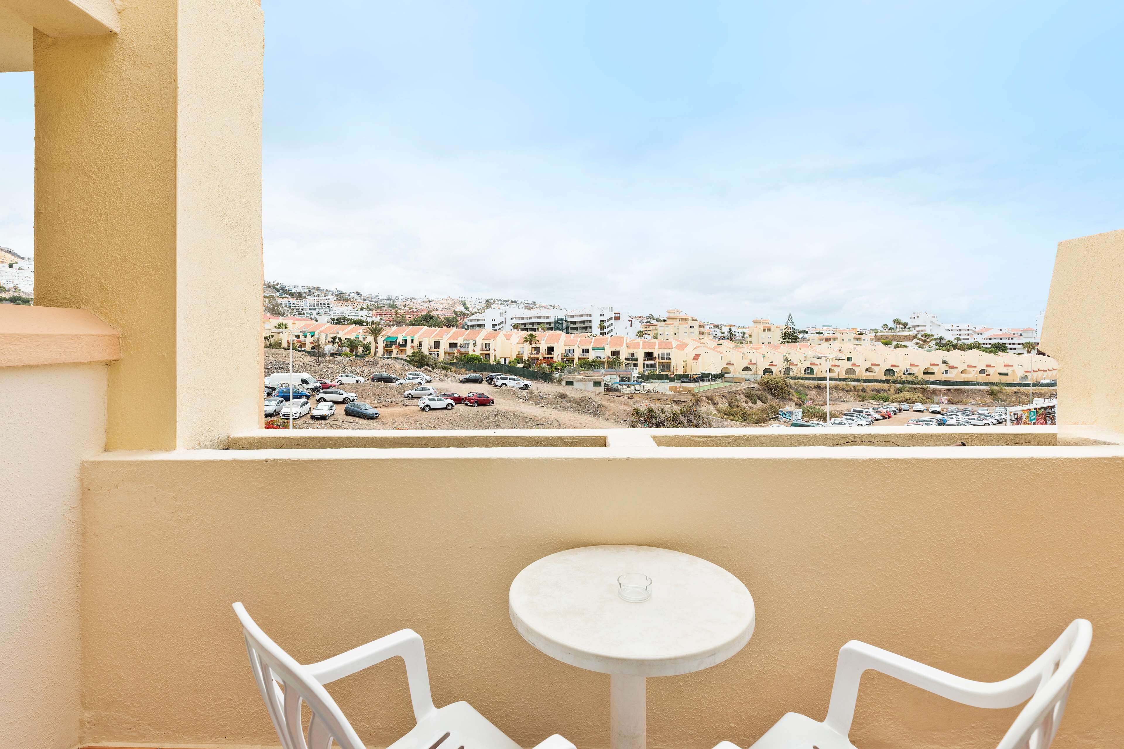 A balcony with a table and two chairs, overlooking a parking area and buildings in the distance.