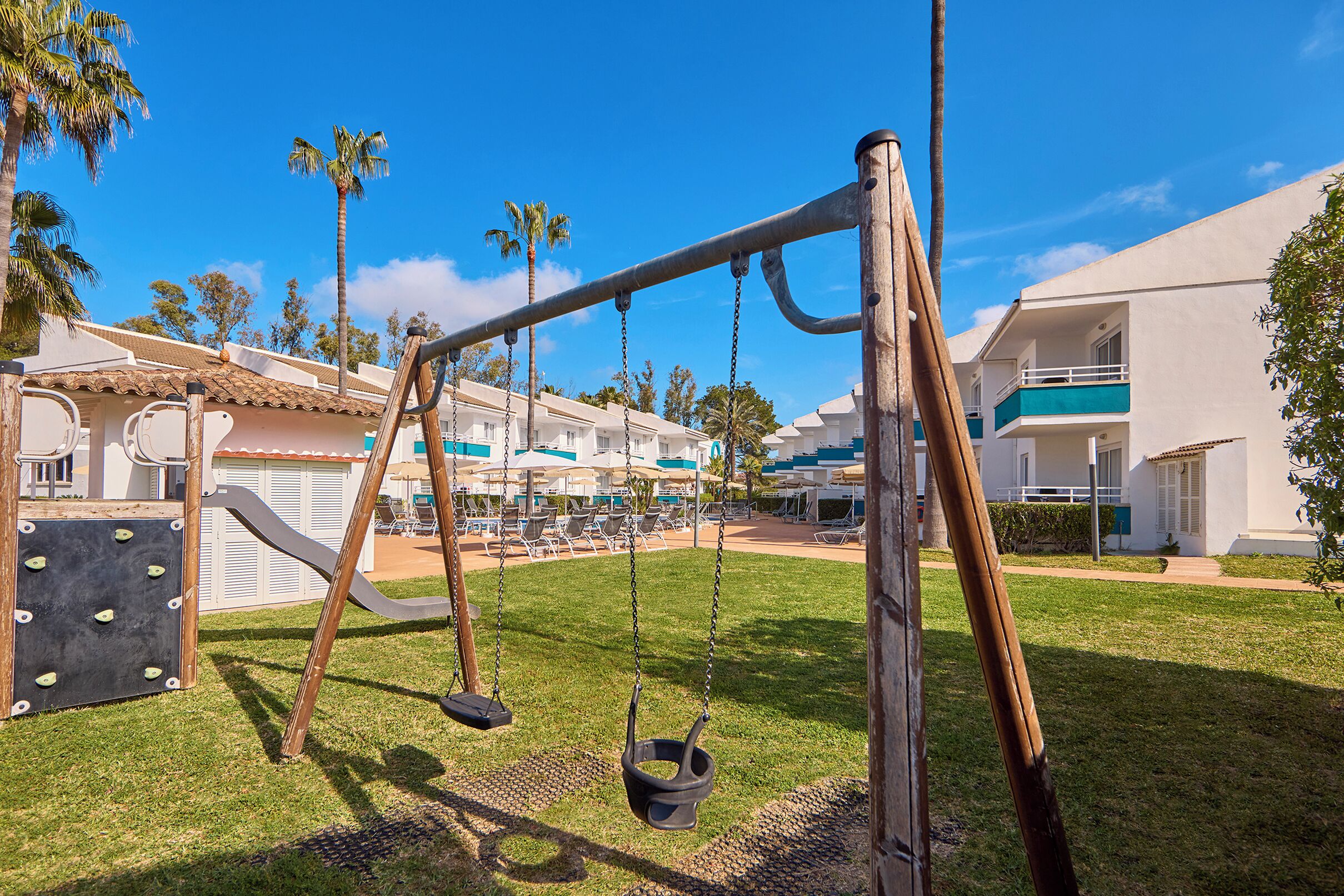 Un parque infantil con columpios y tobogán, rodeado de palmeras y una zona de piscina.