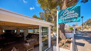Un espace extérieur aménagé avec des meubles en osier et une structure en verre, une enseigne indiquant le restaurant Elisa Lakeside, et un arbre en arrière-plan.