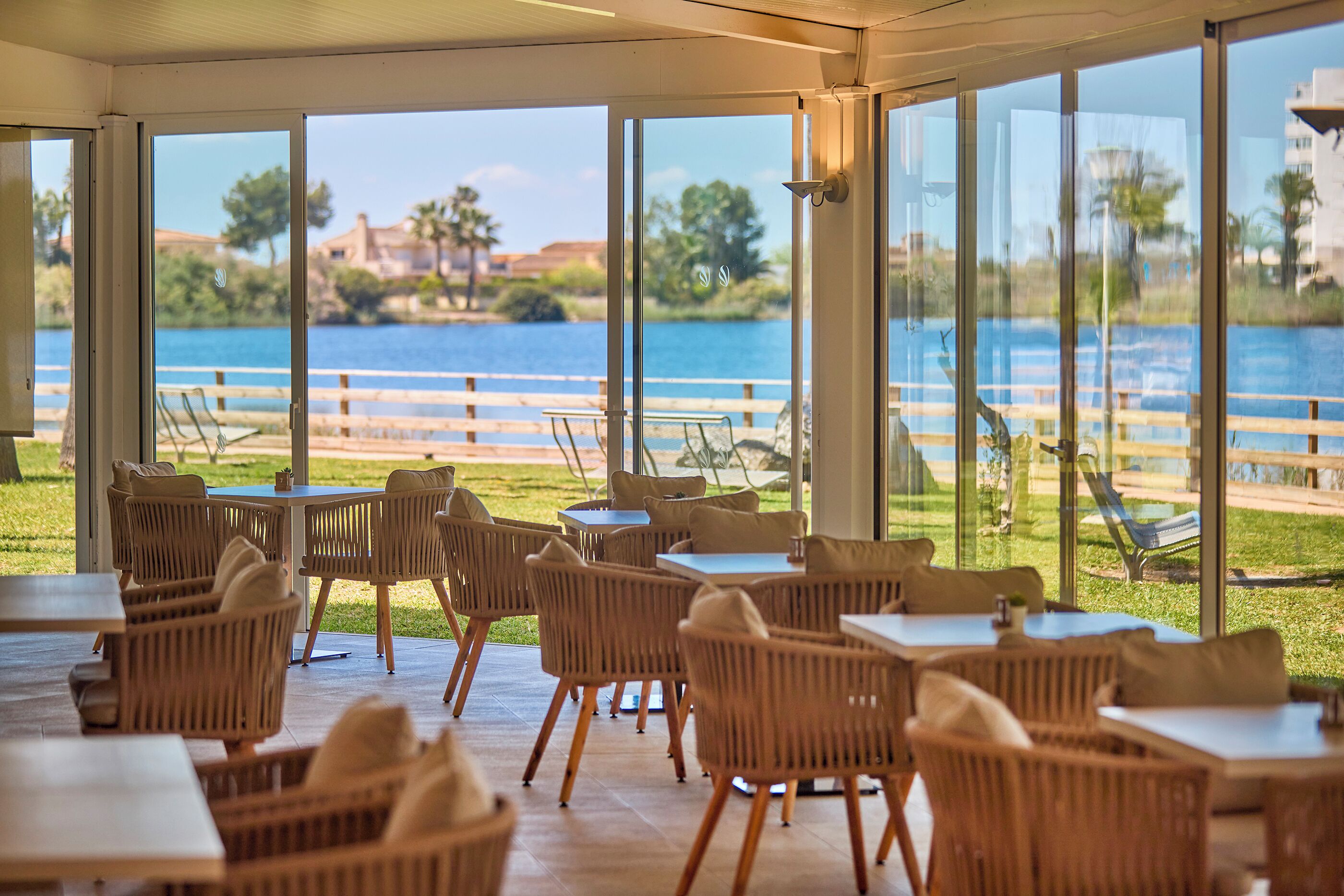 Un espacio de comedor al aire libre techado, con mobiliario de mimbre y ventanas amplias con vistas a un cuerpo de agua y una zona verde.