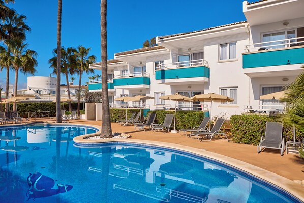 A swimming pool with lounge chairs and umbrellas, surrounded by palm trees and a multi-story building with balconies. - Universal Aparthotel Elisa (Muro)