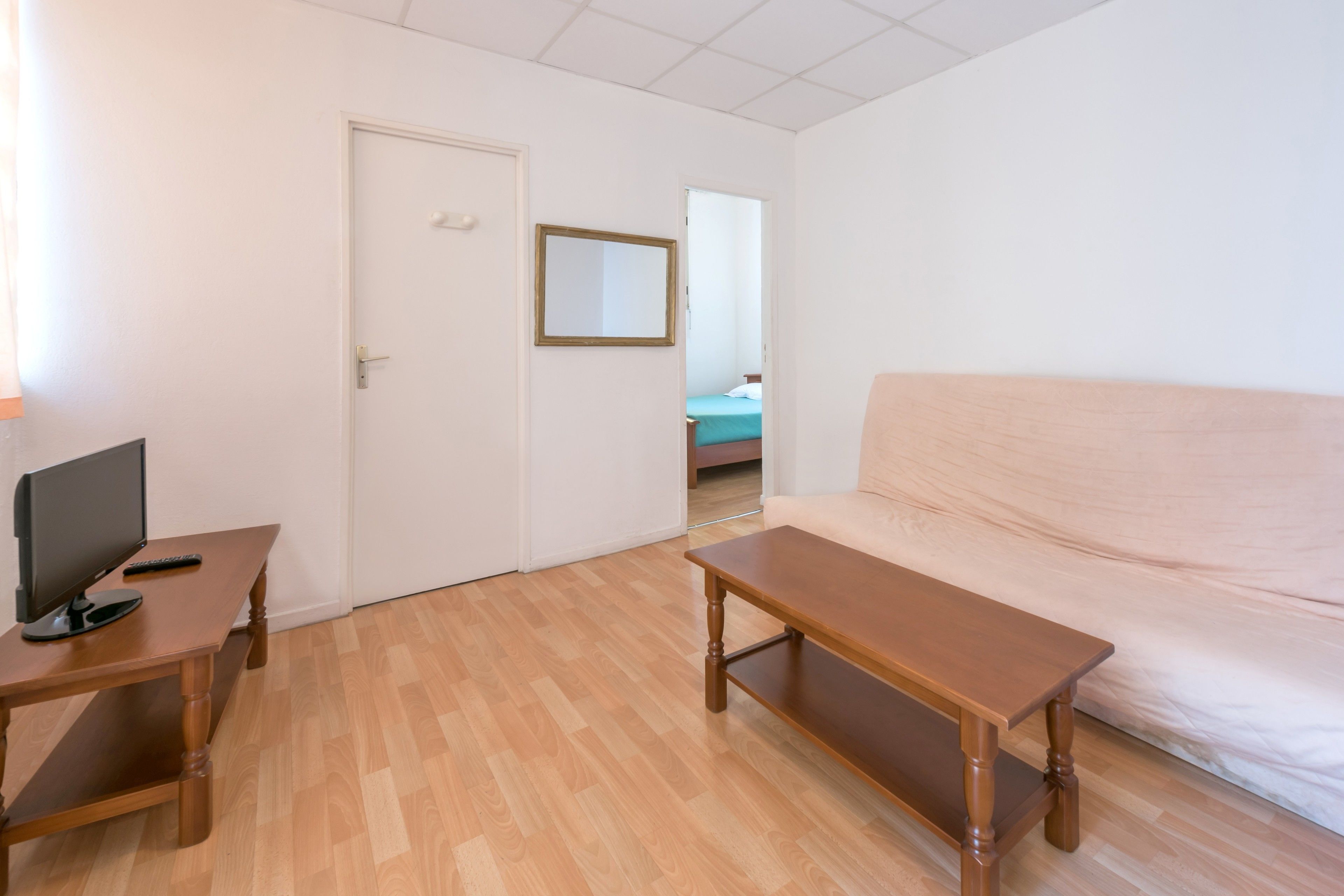 Una habitación con piso de madera, una puerta blanca, una cama con cubrecama azul, un banco de madera y un televisor sobre una mesa de madera.