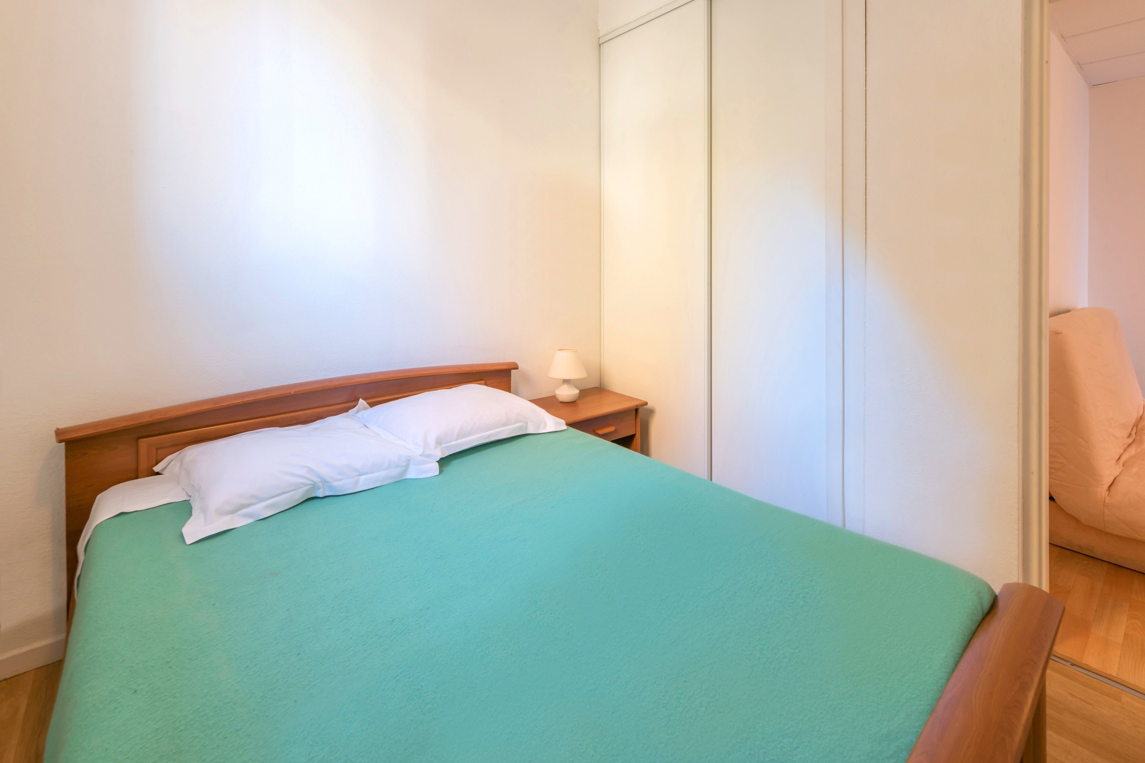 Ein Einzelbett mit grüner Bettdecke und weißen Kissen, ein Nachttisch aus Holz mit einer Lampe sowie ein Kleiderschrank mit Schiebetüren.