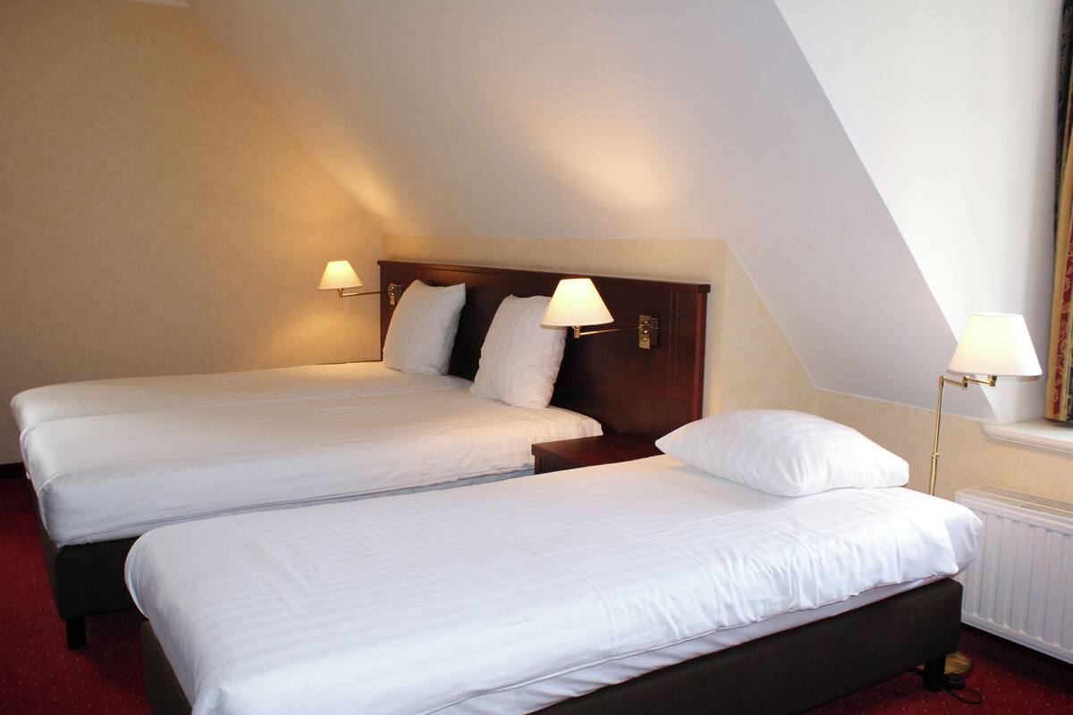 Une chambre d’hôtel avec deux lits simples, chacun doté d’une tête de lit en bois et de linge de lit blanc. Deux lampes murales sont fixées de chaque côté des lits.