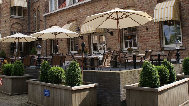 Een terras met stenen muren, houten plantenbakken met struiken en meerdere tafels en stoelen onder gestreepte luifels.