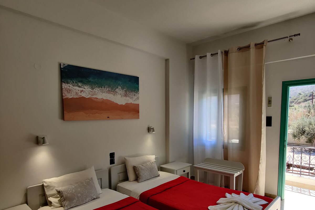 A hotel room with two beds, a painting of a beach scene, a window with curtains, and a view of the outdoors.