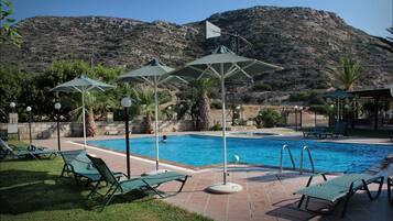 Zona de piscina con sillas reclinables y sombrillas, rodeada de vegetación y un telón de fondo montañoso.