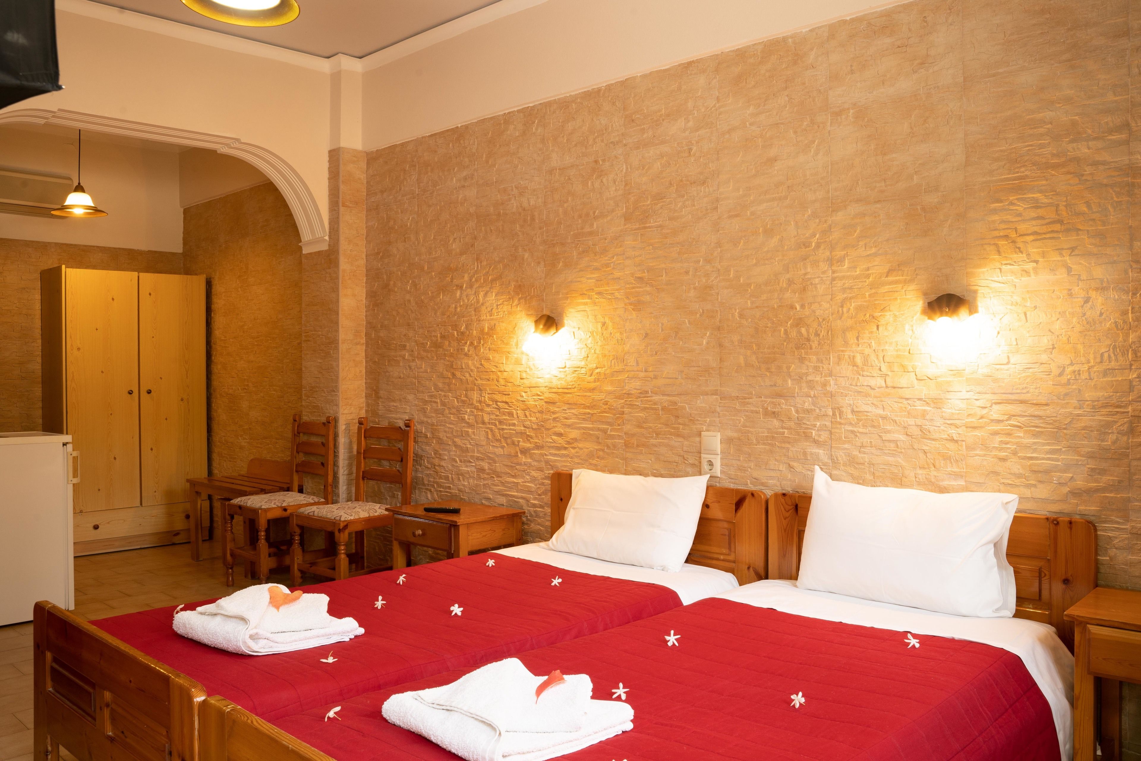 Habitación de hotel con dos camas, cabeceros de madera y una pared de piedra. Hay dos mesitas de noche, un armario de madera y un escritorio con sillas.