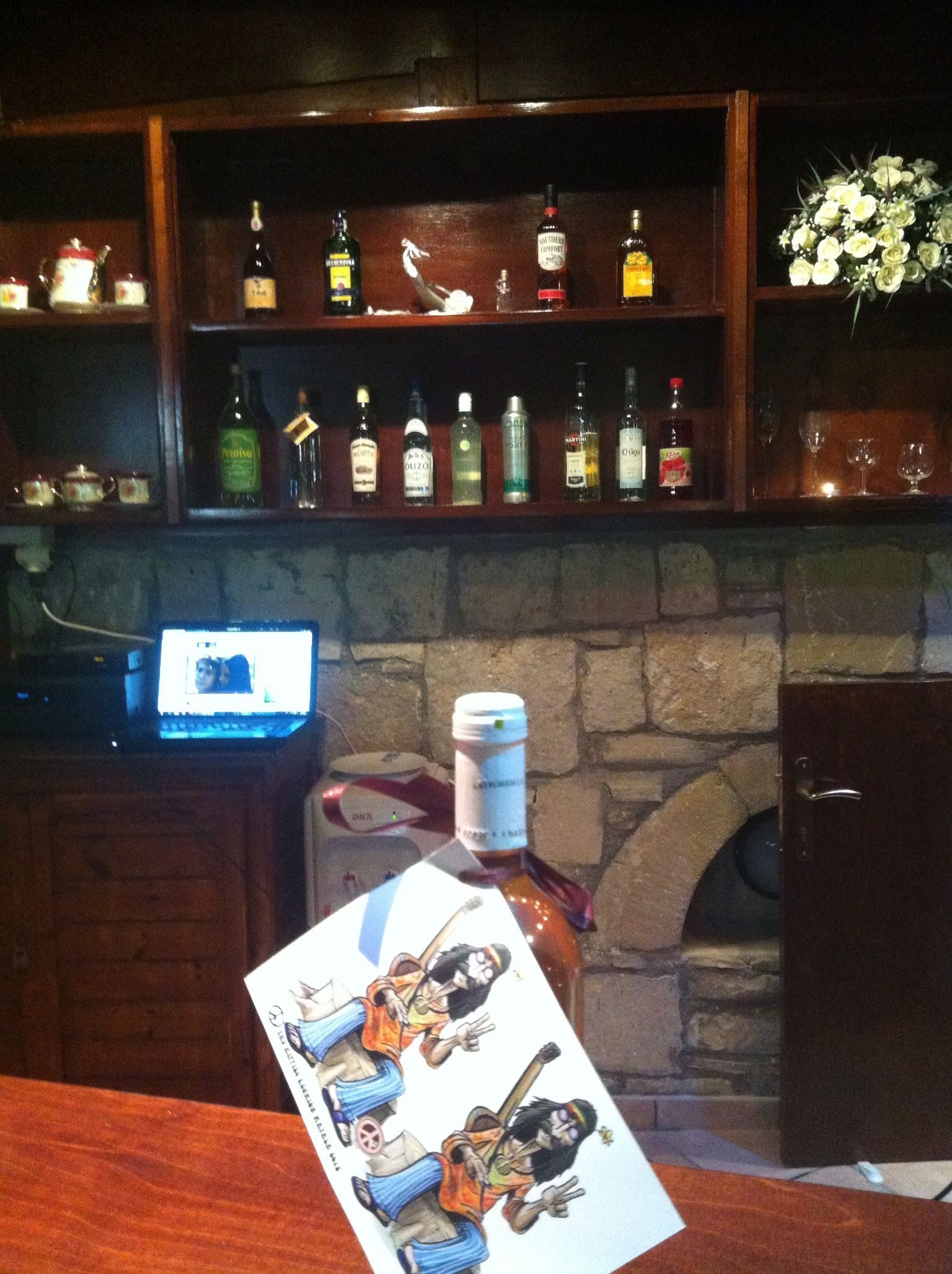 A bar area with shelves of liquor bottles, a laptop on a table, and a decorative card.
