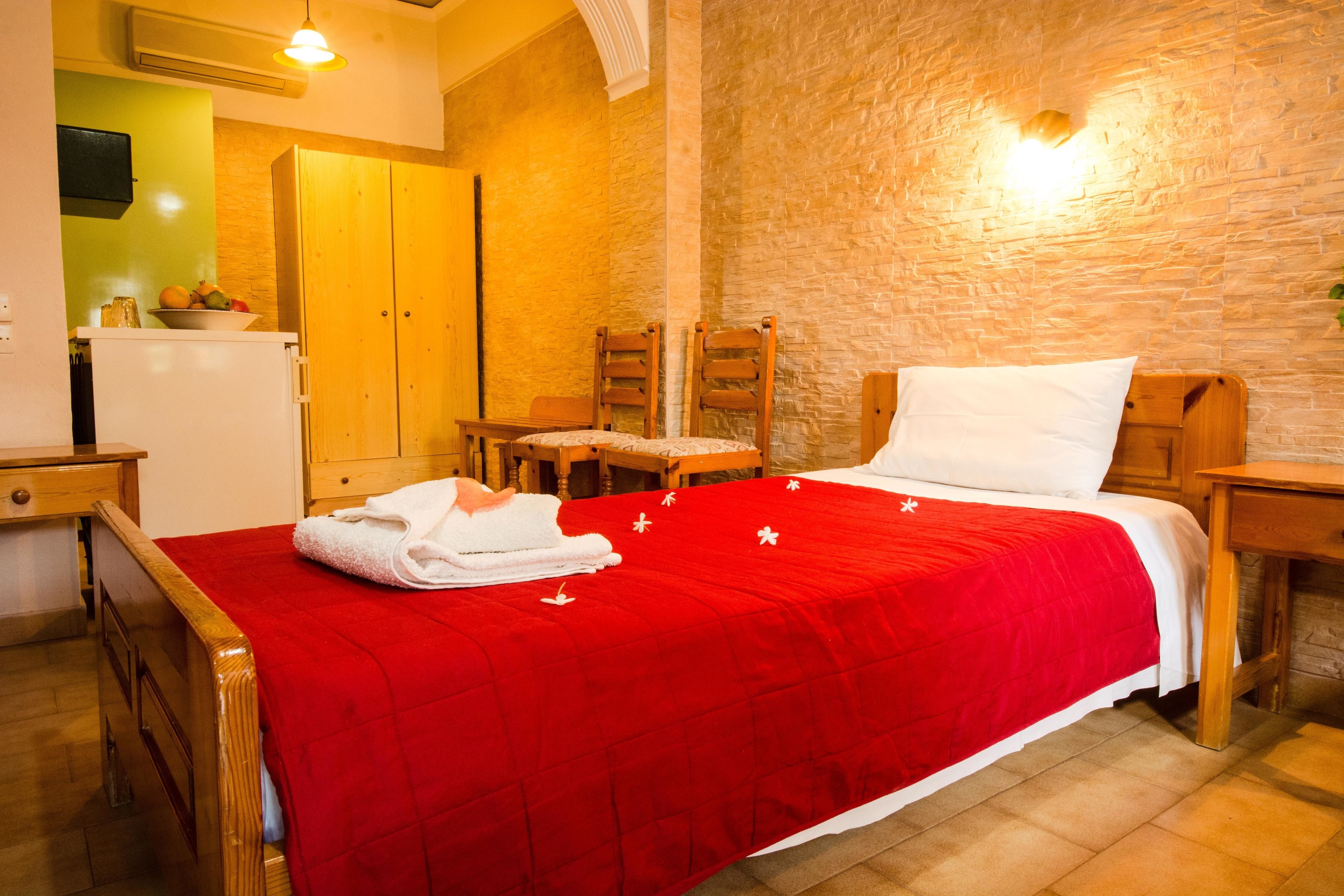 A hotel room with a red bedspread, wooden furniture, a small table, a chair, and a wall-mounted light fixture.