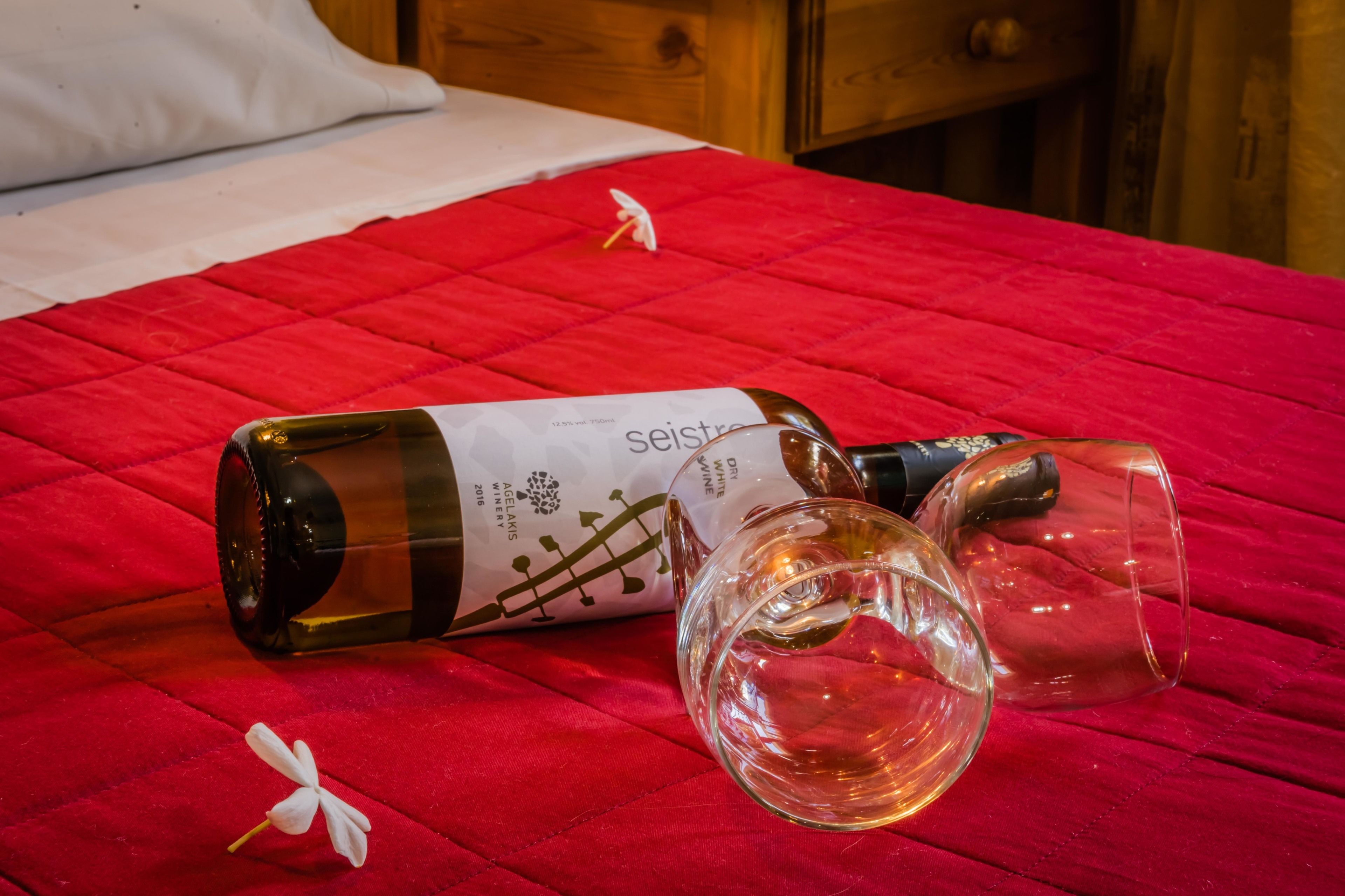 A bed with a red quilt, a bottle of Sejstrup wine, and two empty wine glasses on it.