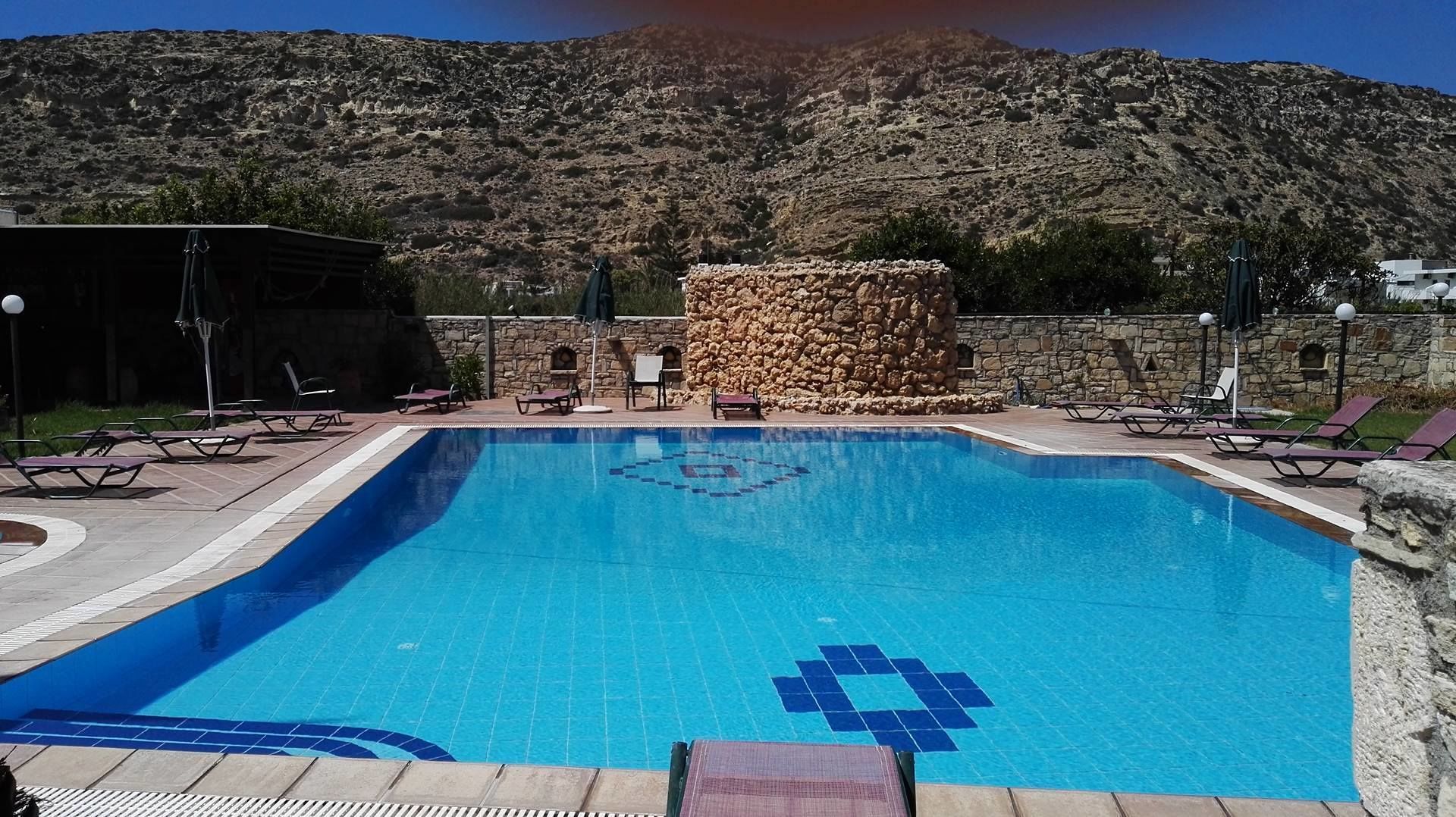 Una piscina rectangular con baldosas azules y un diseño geométrico en el centro, rodeada de sillas de descanso y un muro de piedra.
