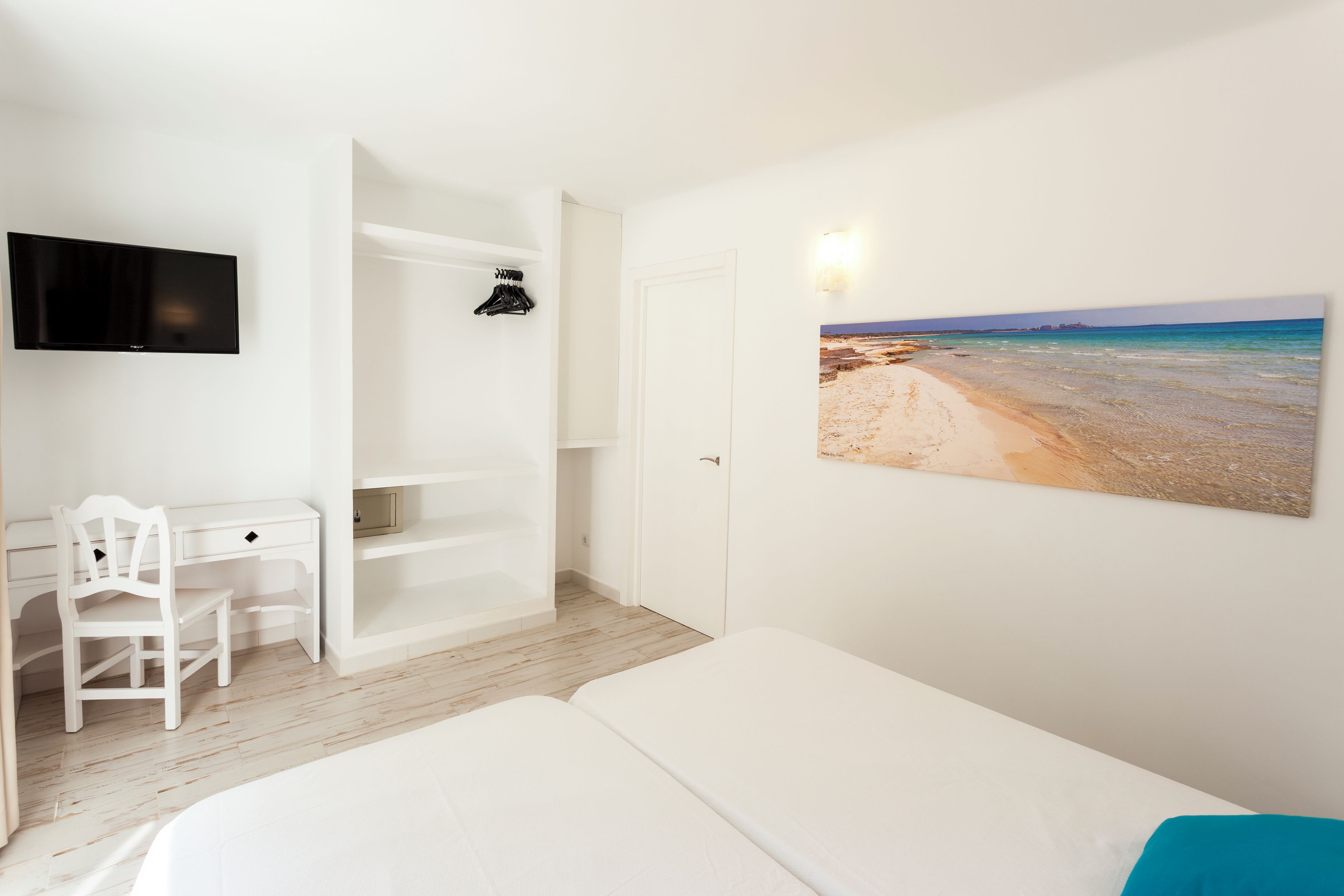 Een slaapkamer met een bed, bureau, stoel, kledingkast, televisie en een schilderij van een strand.