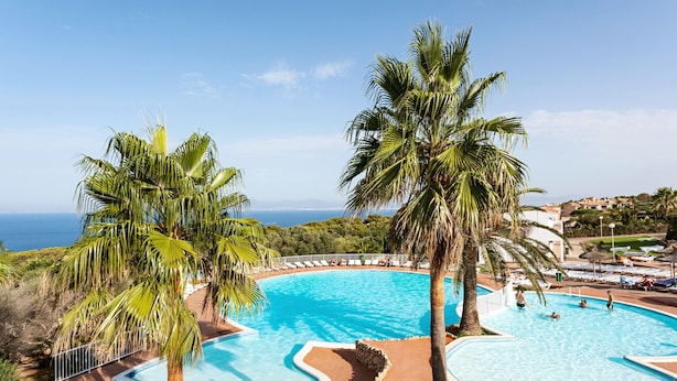 A large, clear swimming pool with a curved edge, surrounded by palm trees and a view of the sea. - Sun Club El Dorado (Llucmajor)