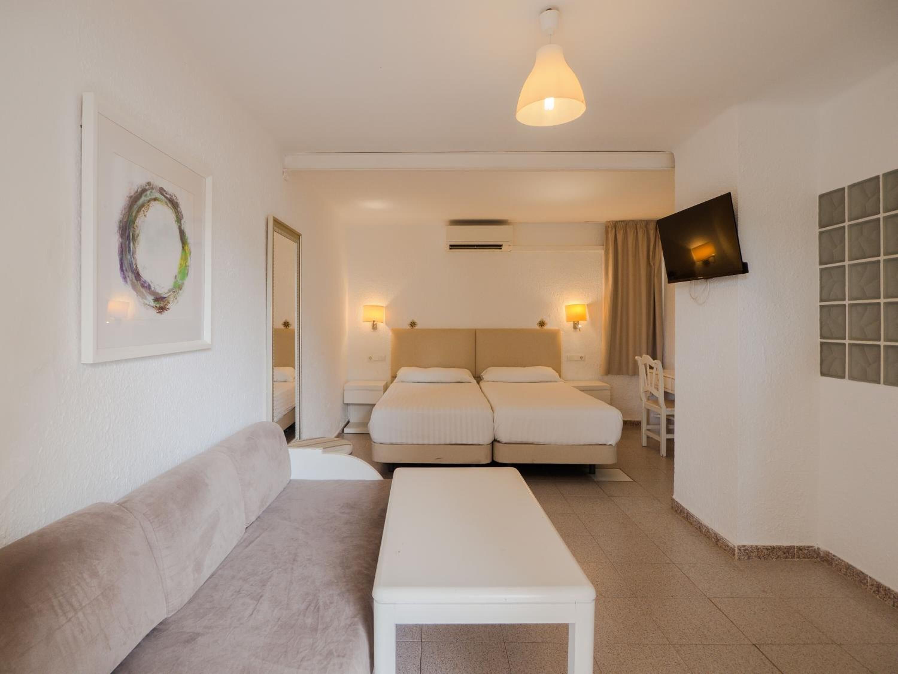 A hotel room with a sofa, a small table, two beds, a TV, and a wall-mounted air conditioning unit.
