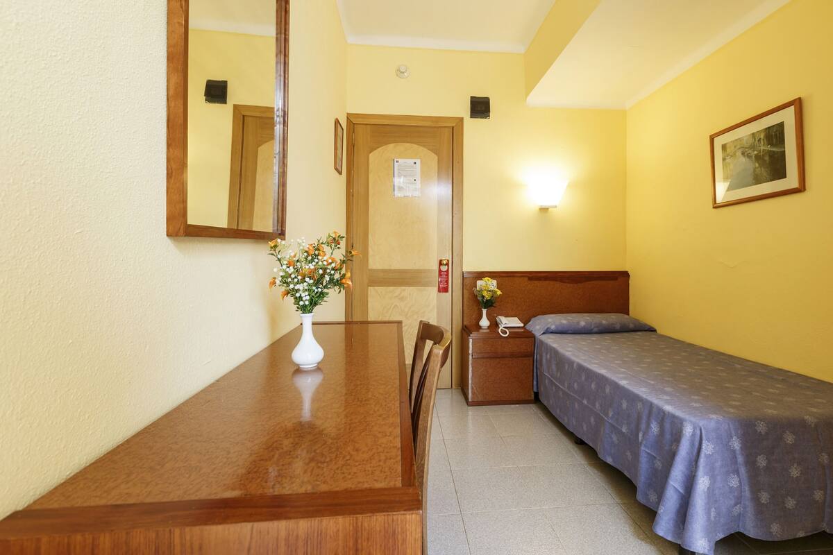Una habitación de hotel pequeña con una mesa de comedor de madera, una cama individual con colcha azul, una mesita de noche con lámpara y un espejo en la pared.