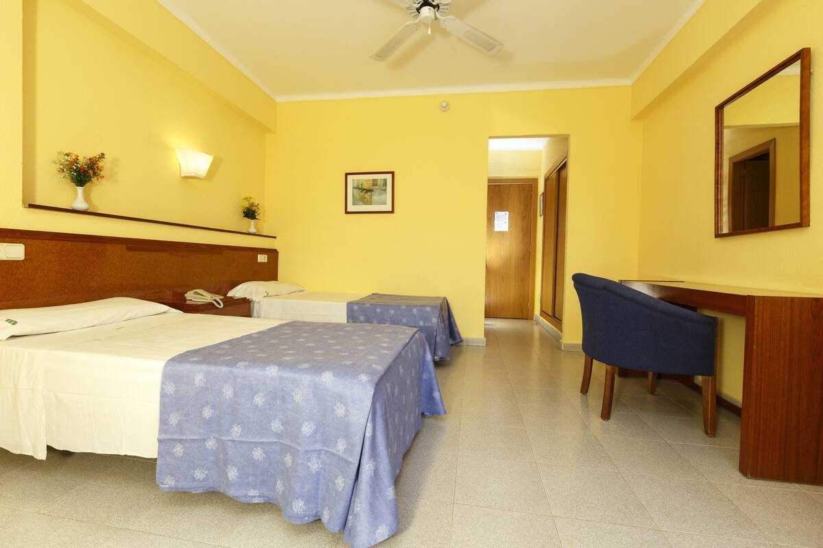 Habitación de hotel con cama, dos mesitas de noche, escritorio, silla, ventilador de techo y una puerta que da a otra habitación.