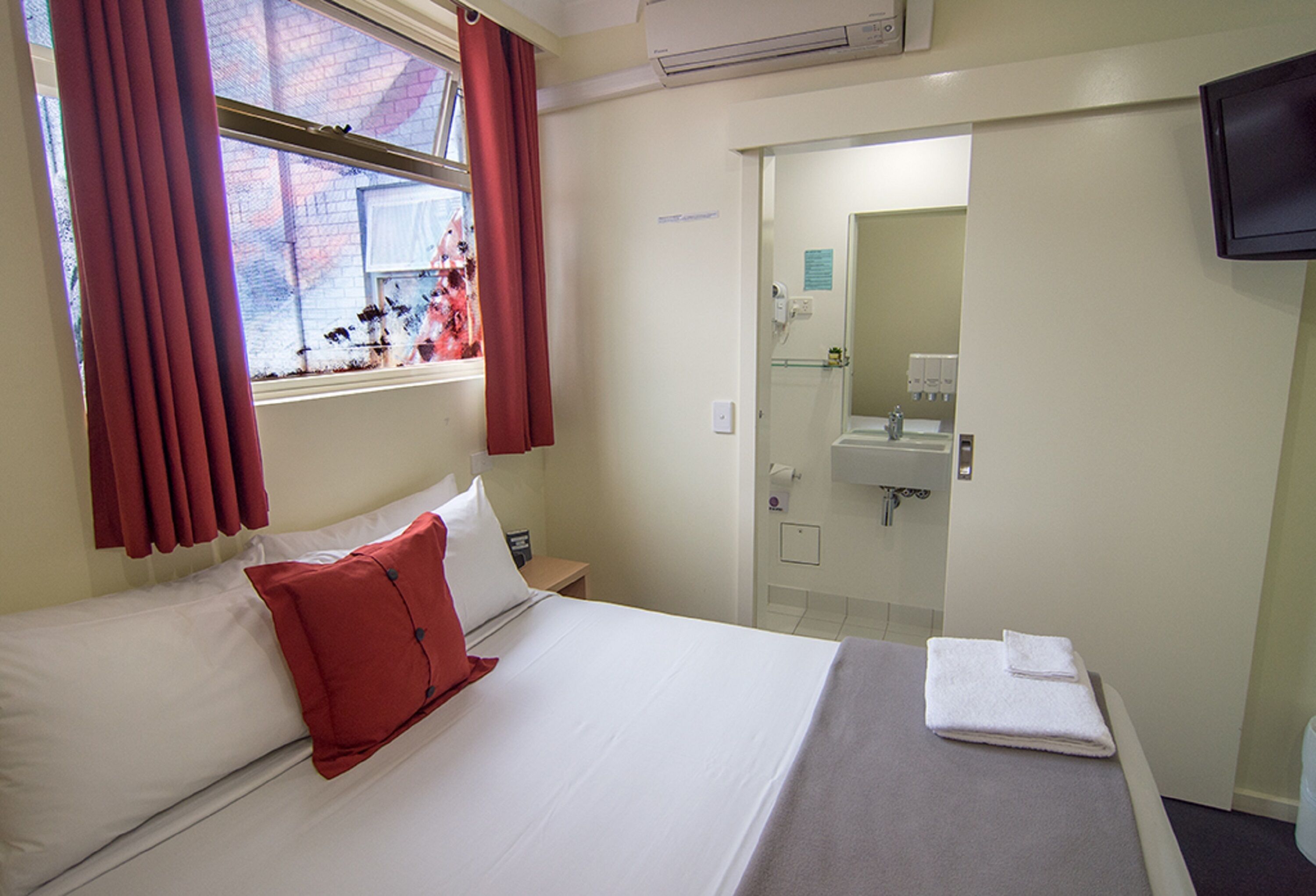 A hotel room with a bed, a window with red curtains, a bathroom with a sink and mirror, and a flat-screen TV mounted on the wall.