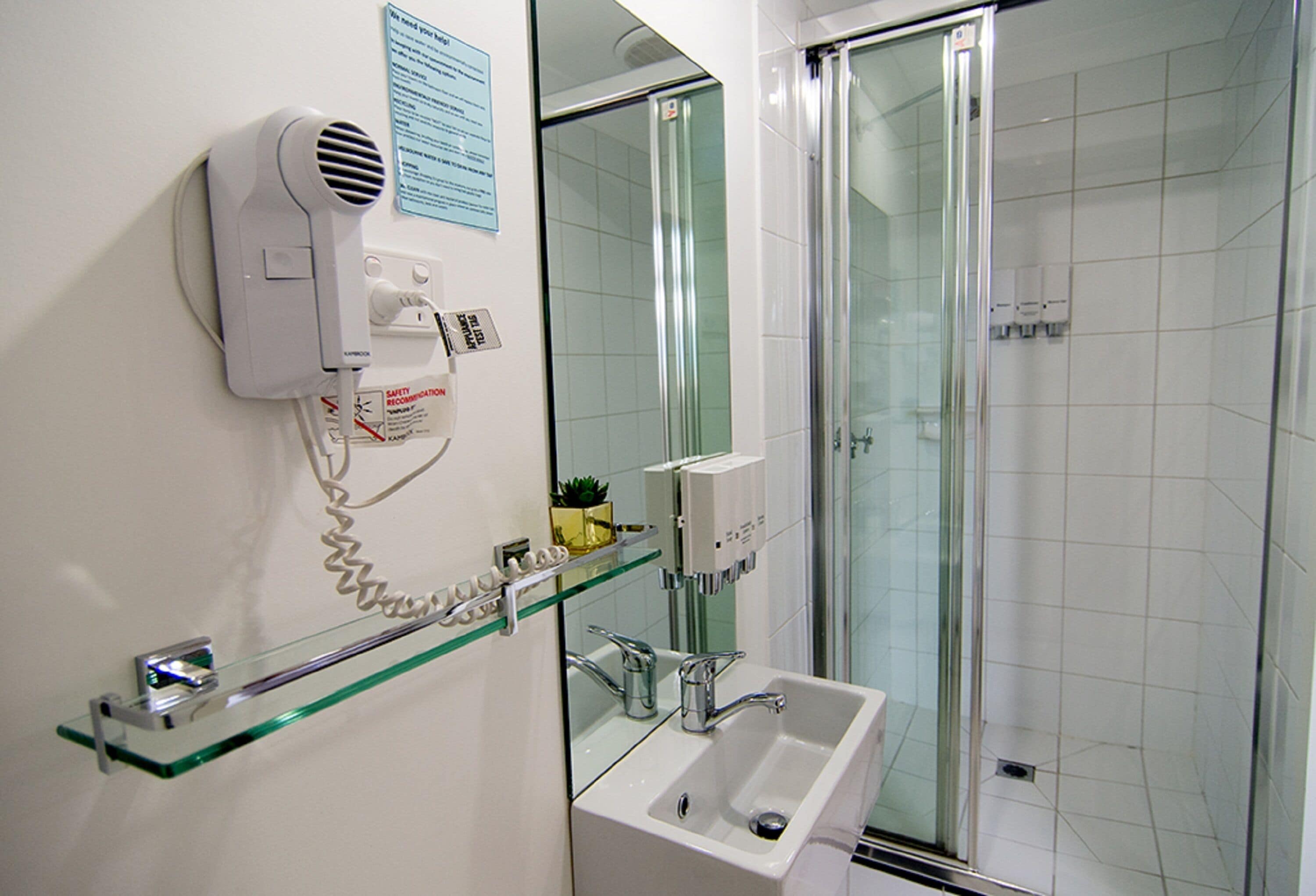Une salle de bain avec une douche à parois de verre, un lavabo blanc, une étagère en verre où sont rangés les articles de toilette et un sèche-cheveux fixé au mur.