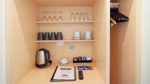 Habitación compacta con kitchenette, que incluye tetera, cafetera y juego de té. Se observan estantes con copas de vino, tazas y un control remoto.