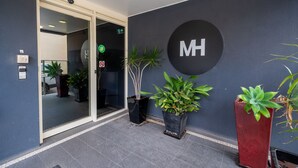 Entrada moderna de un hotel con puerta de cristal, plantas en macetas y un gran cartel con la inicial 'MH' en la pared.