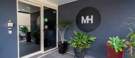 A modern hotel entrance with a glass door, potted plants, and a large 'MH' sign on the wall.