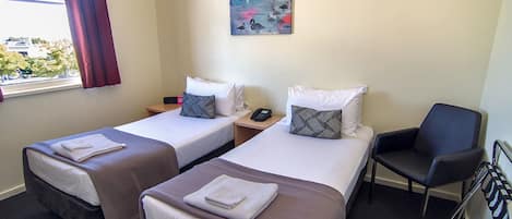 A hotel room with two single beds, a chair, a painting on the wall, and a window with curtains.