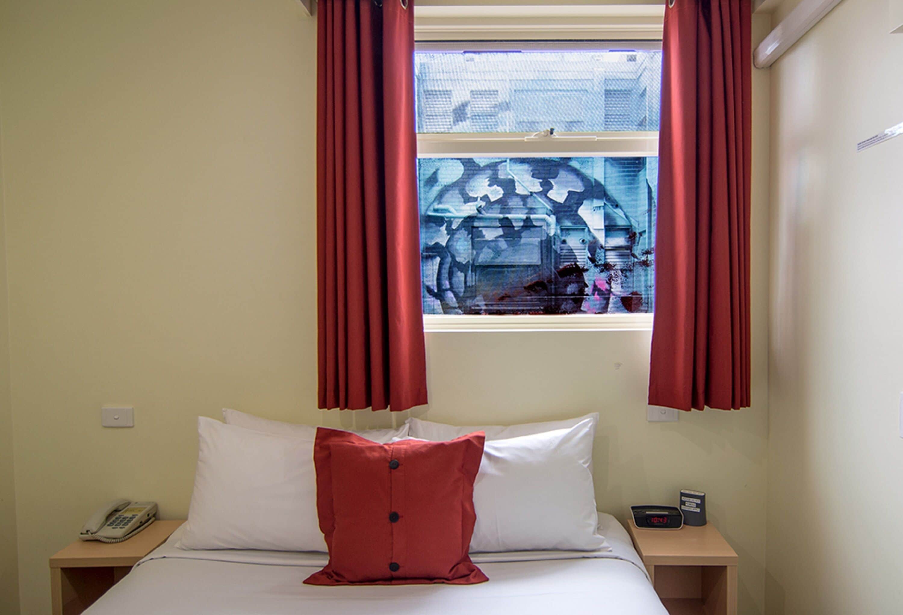 Una habitación de hotel con una cama, una mesita de noche con un teléfono, un reloj digital y una ventana con cortinas rojas.