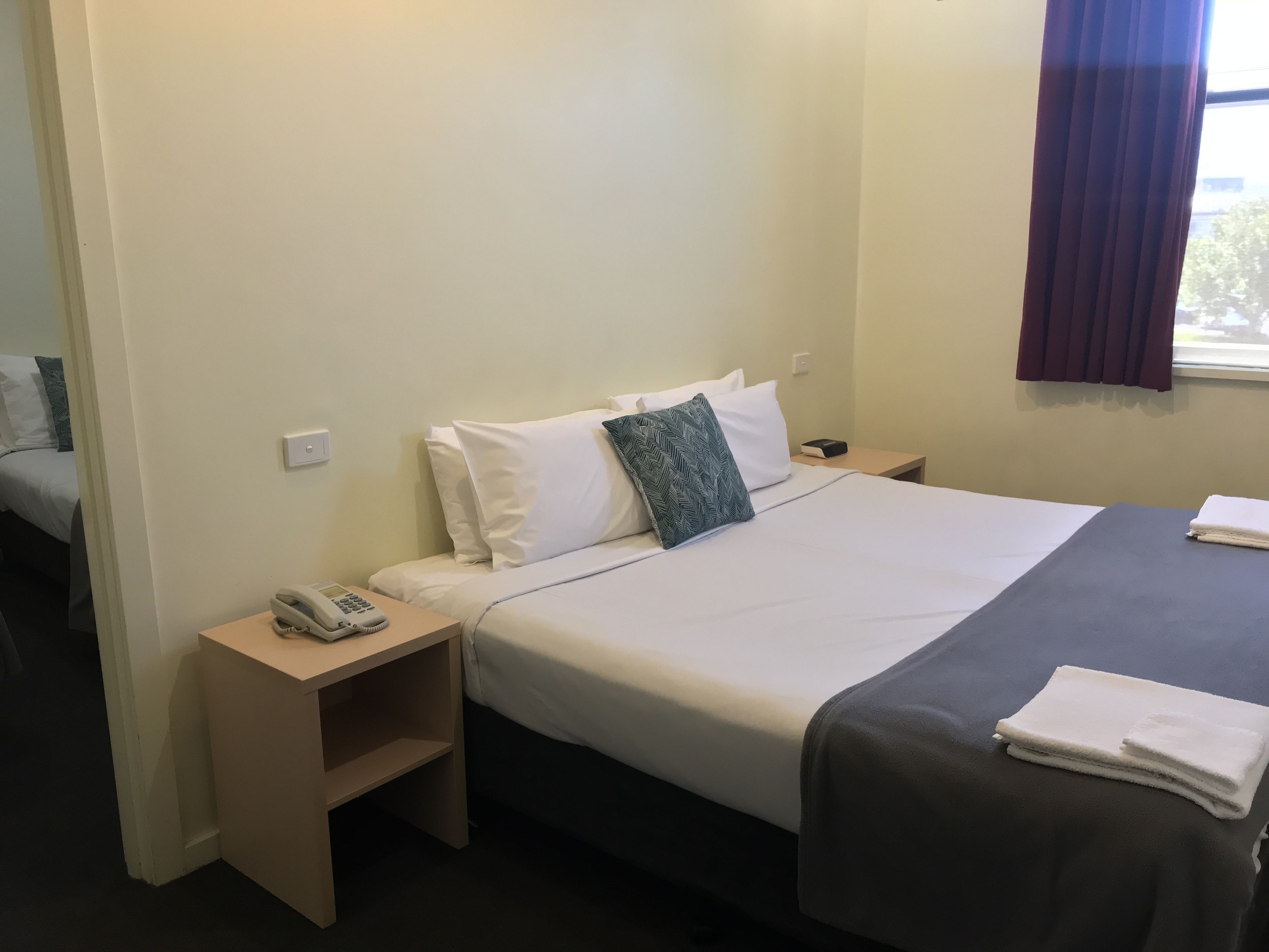 Et hotelværelse med en seng, et natbord med telefon, en sengebord og et vindue med gardiner.
