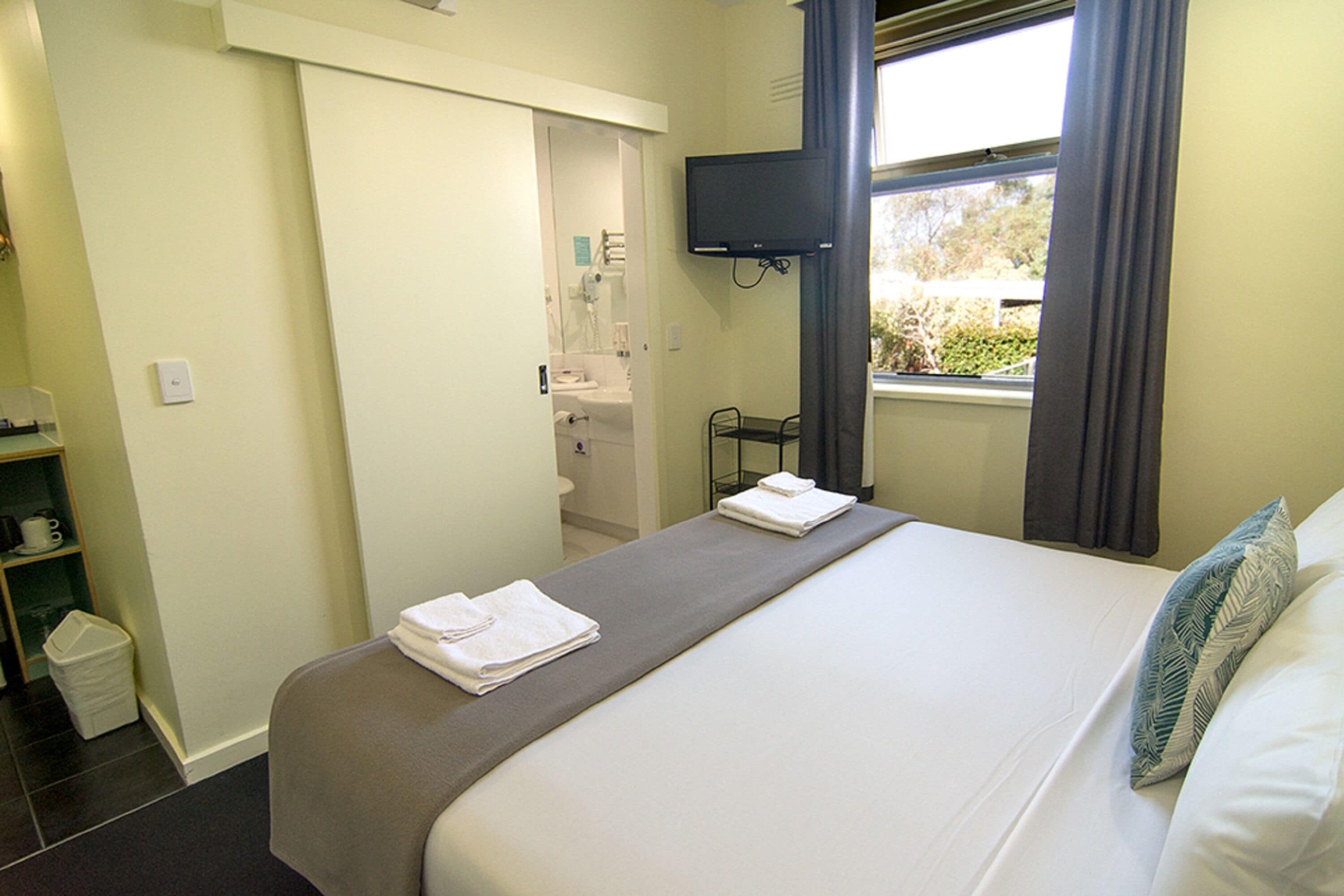 A hotel room with a bed, a TV mounted on the wall, a window with curtains, and a bathroom visible through an open door.