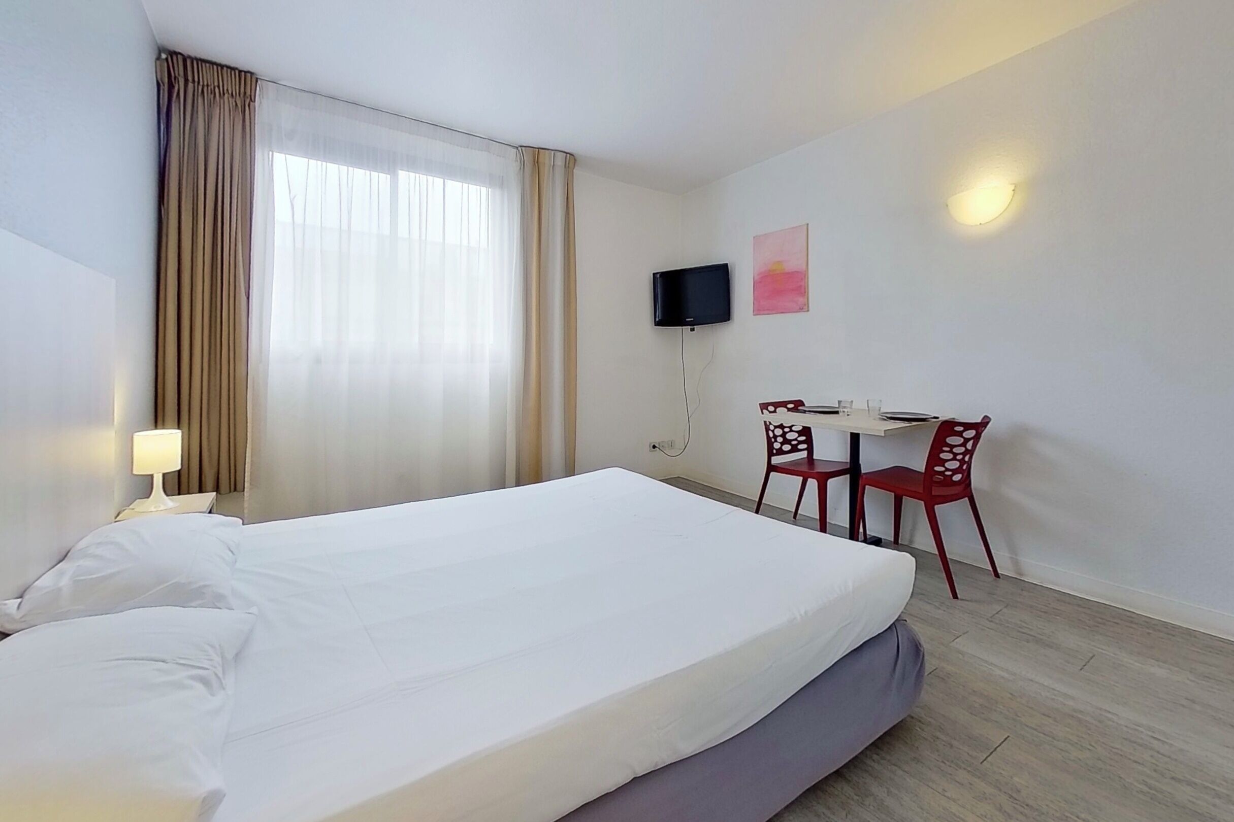 A hotel room with a large bed, a desk with two chairs, a TV mounted on the wall, and a window with curtains.