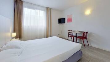 A hotel room with a large bed, a desk with two chairs, a TV mounted on the wall, and a window with curtains.