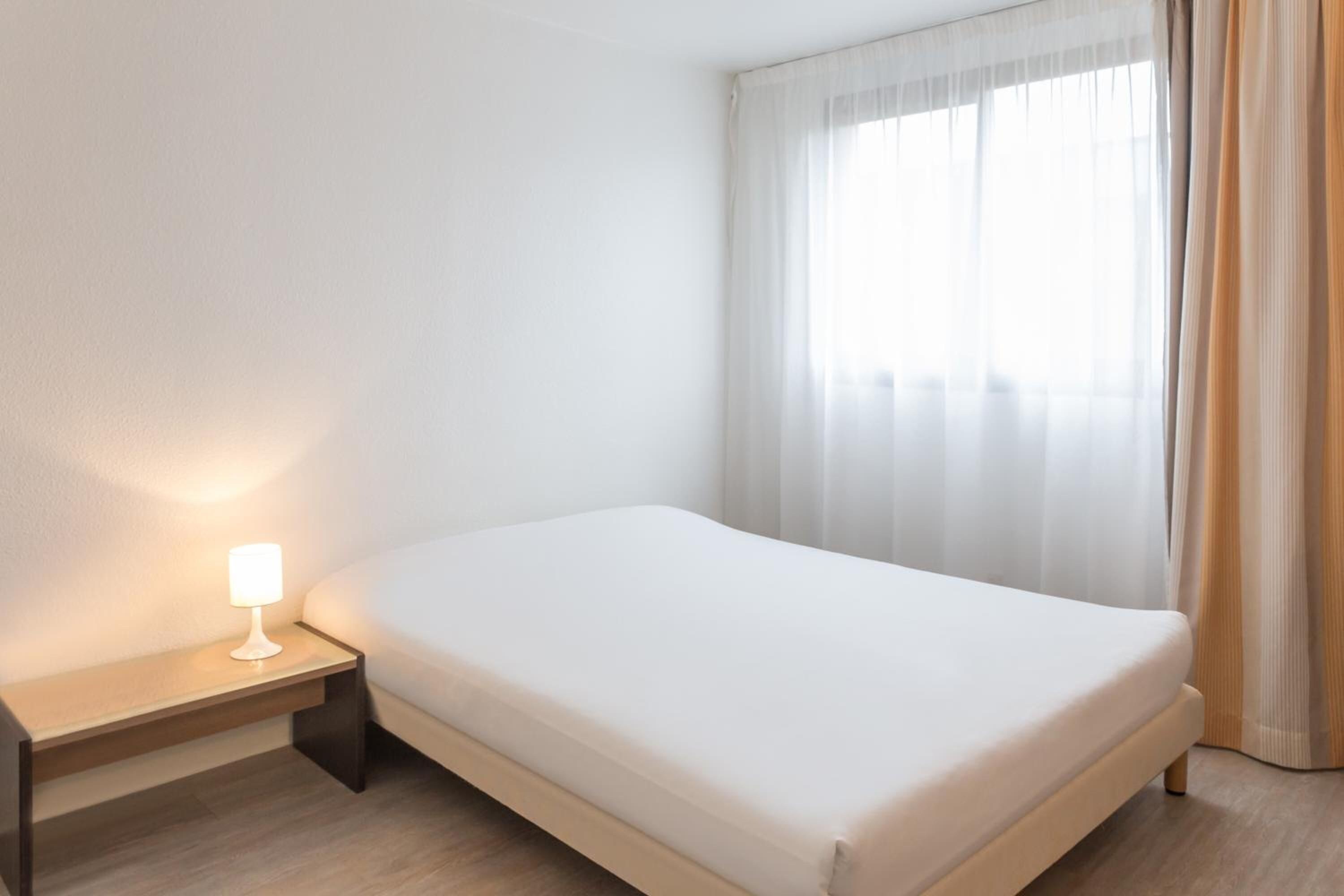 A hotel room with a bed, a bedside table with a lamp, and a window with curtains.