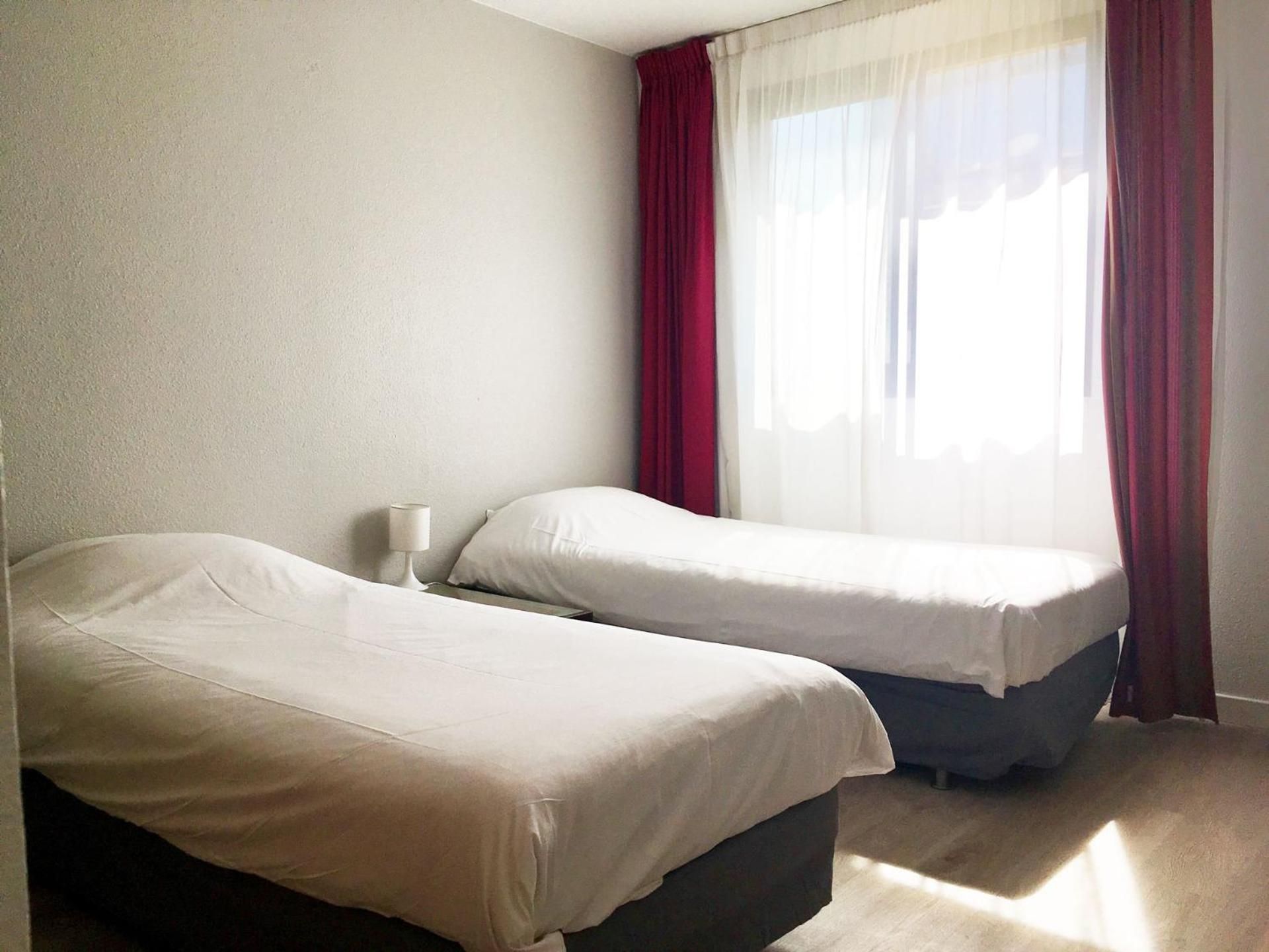 A hotel room with two beds, a bedside table, and a window with sheer curtains.
