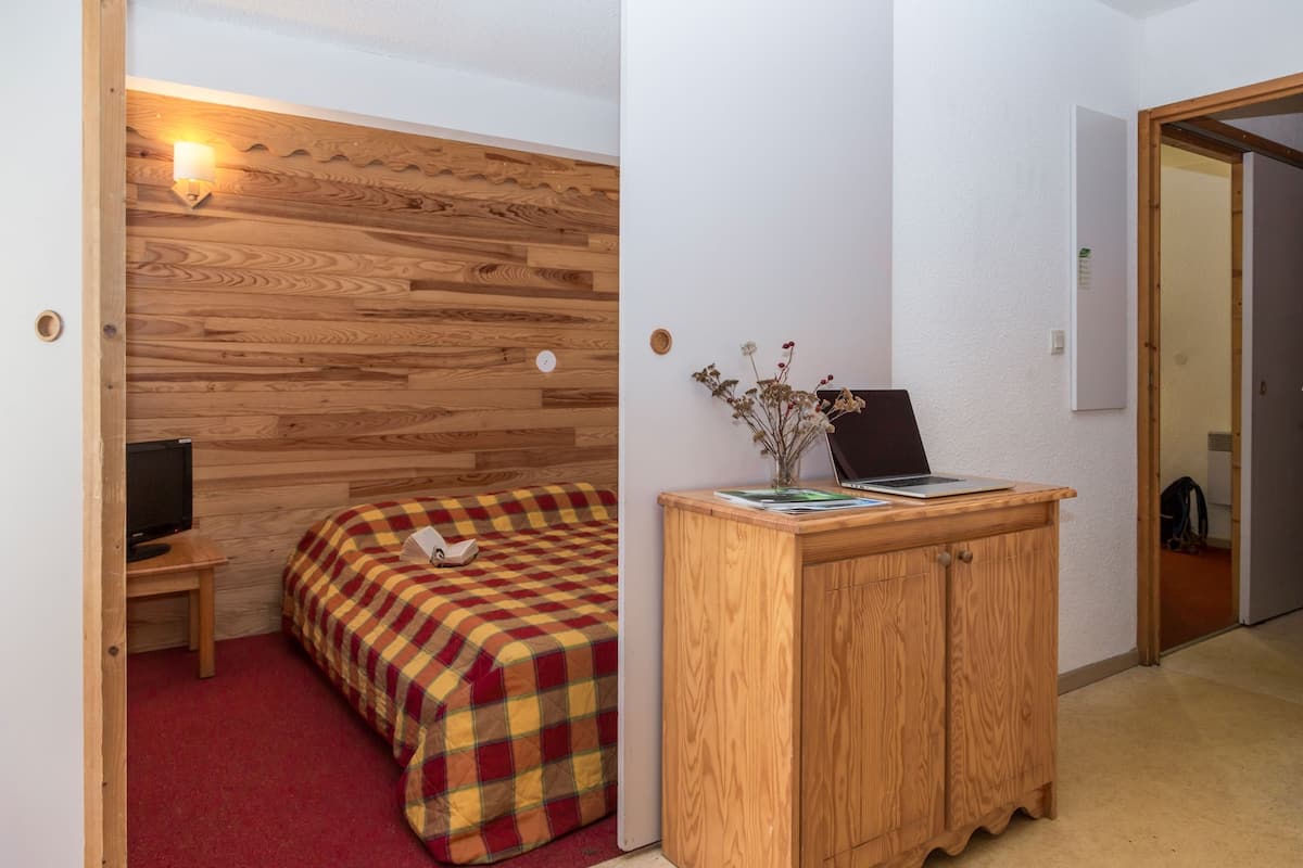 Una habitación con pared de madera, una cama con manta de cuadros, un escritorio con una computadora portátil y un pequeño armario con un jarrón de flores.