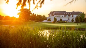 A building with a flat roof and multiple windows, surrounded by a well-maintained lawn and a small pond. - Greenfield Hotel Golf & Spa (Buk)