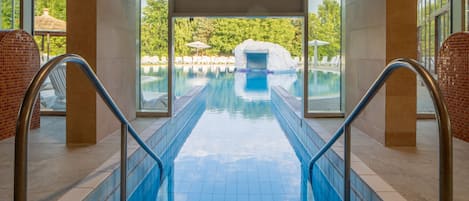 Una piscina interior con paredes de vidrio transparente, piso de baldosas y vistas a un área exterior con sombrillas y sillas de descanso.