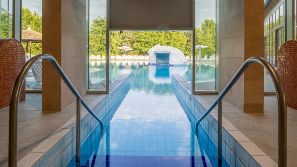 An indoor swimming pool with clear glass walls, a tiled floor, and a view of an outdoor area with umbrellas and lounge chairs. - Greenfield Hotel Golf & Spa (Buk)