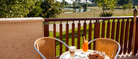 A round table with wicker chairs, a glass of orange juice, a basket of pastries, and a beverage on a terrace overlooking a lake.