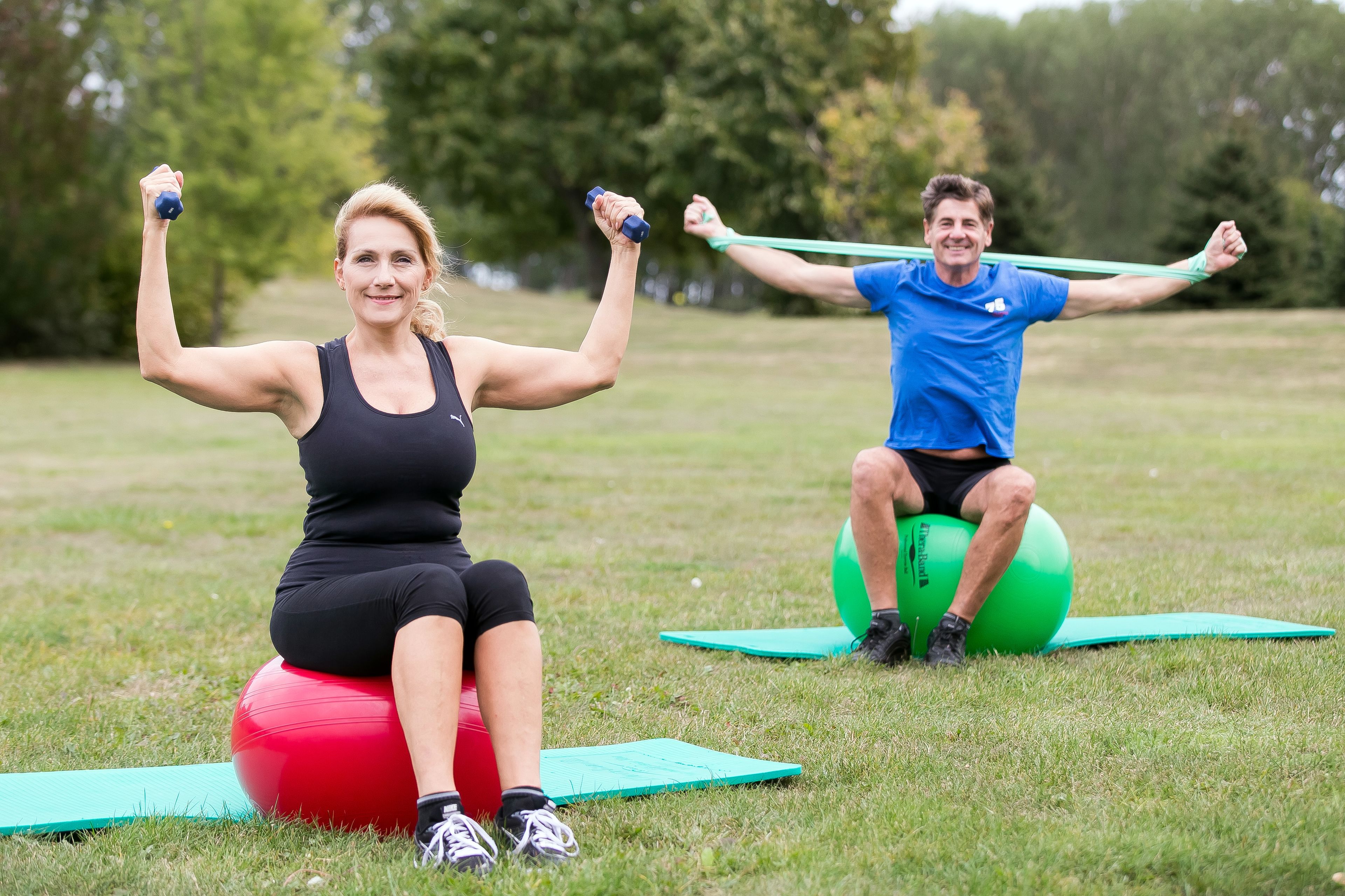 Two people exercising on exercise balls in an outdoor grassy area with trees in the background.