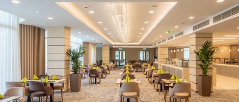 A spacious hotel lobby with multiple dining areas, large windows, and a modern chandelier.