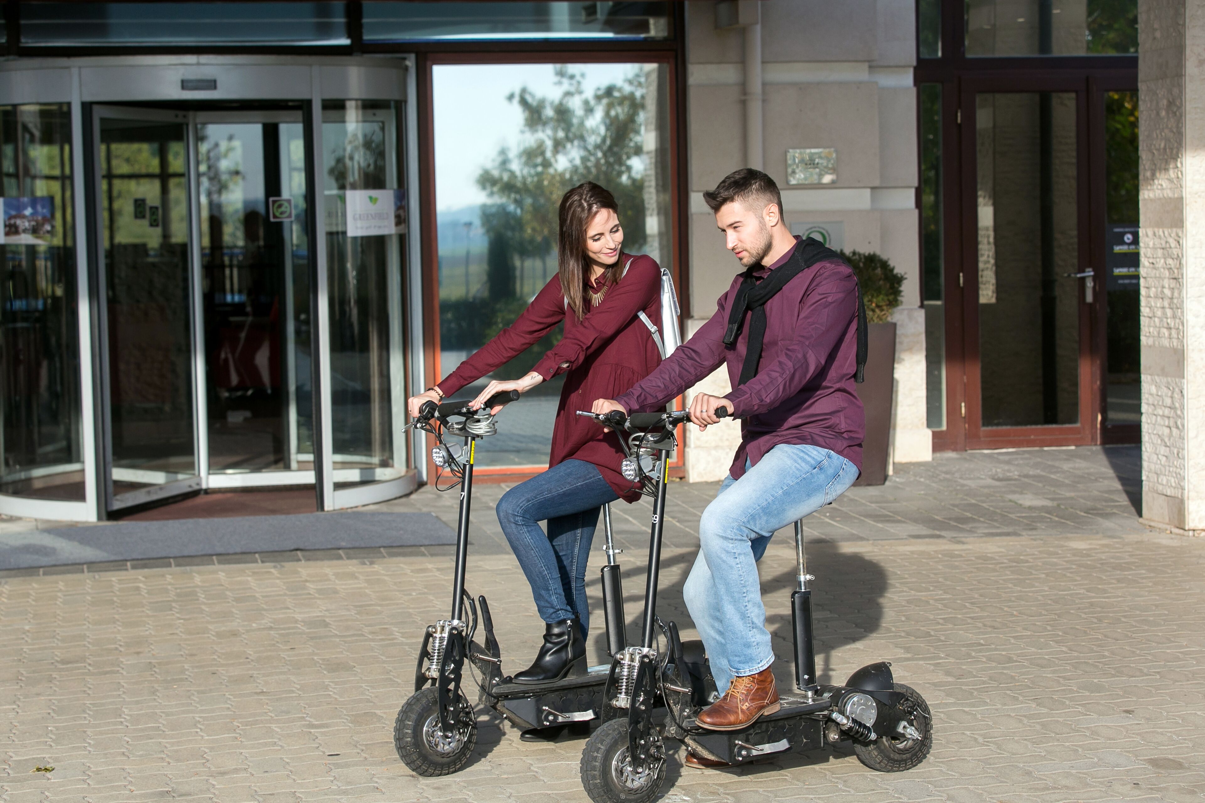 Two people riding electric scooters in front of a building with glass doors and a stone facade.