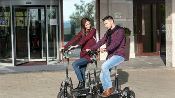 Two people riding electric scooters in front of a building with glass doors and a stone facade.
