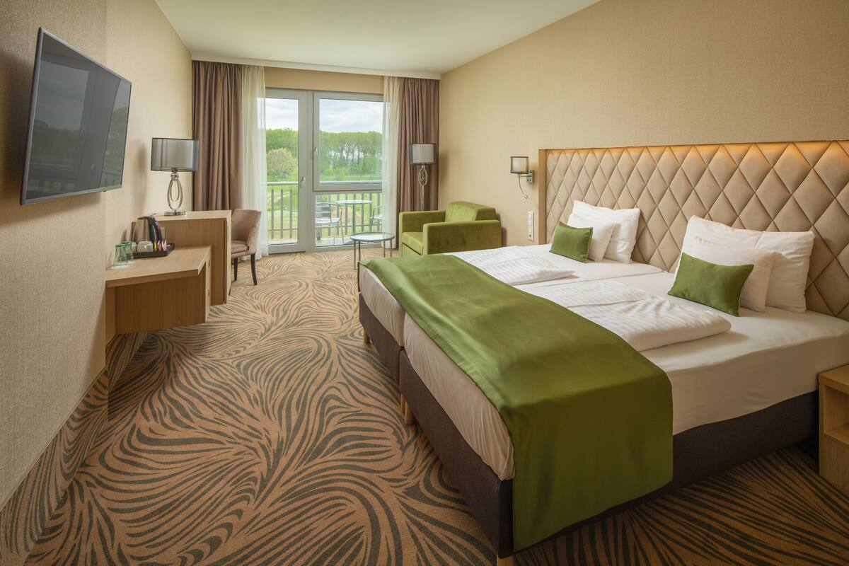 Ein Hotelzimmer mit einem großen Bett, einem Schreibtisch, einem Sessel, einem Fernseher und einem Balkon mit Blick auf Bäume.