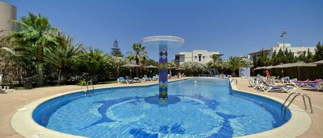 Uma piscina circular com uma fonte decorativa no centro, rodeada por espreguiçadeiras e palmeiras.